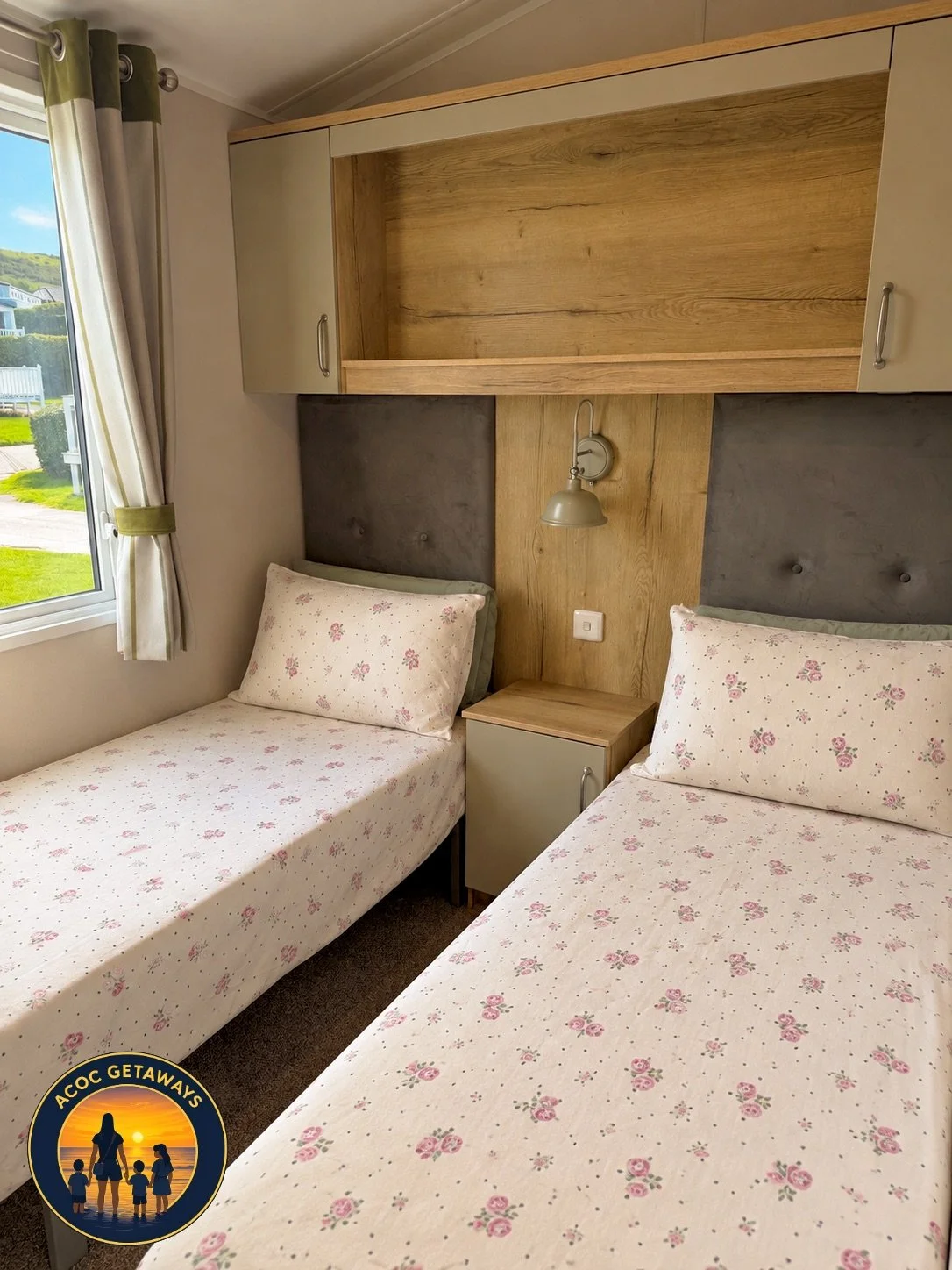 Two single beds with floral bedsheets and pillows, a small nightstand, a wall lamp, a window with curtains, and a built-in wooden shelf in the background.