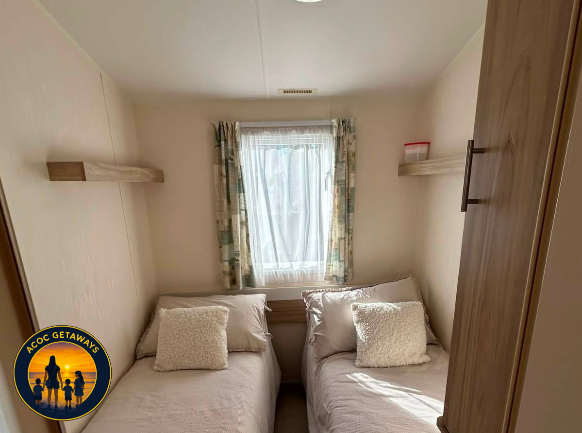 Small bedroom with two single beds with white bedding and pillows, a window with patterned curtains, and wooden shelves on the walls.