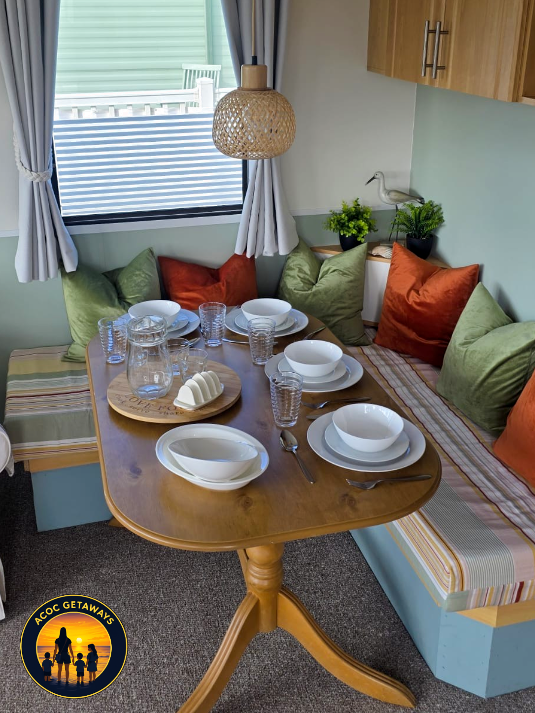 A cozy dining nook with a wooden table set with white bowls, plates, and glasses, surrounded by colorful cushions in a corner near a window decorated with curtains, with potted plants and a wall-mounted decorative bird accent.