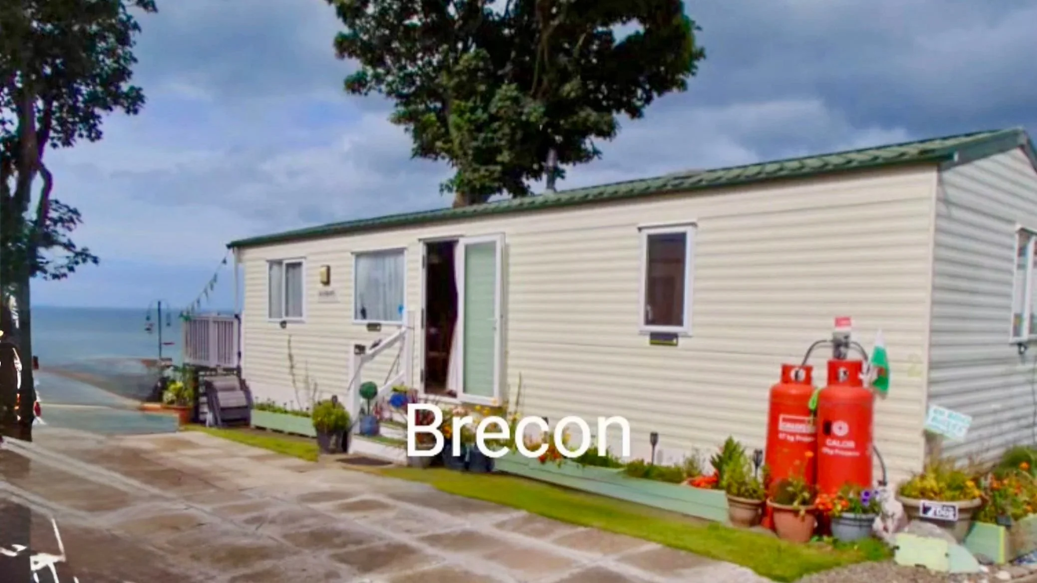 A small, white mobile home with a green roof situated near the beach. It has potted plants and flowers outside, and two red propane tanks attached to the side. The scene is partly cloudy with the ocean visible in the background.