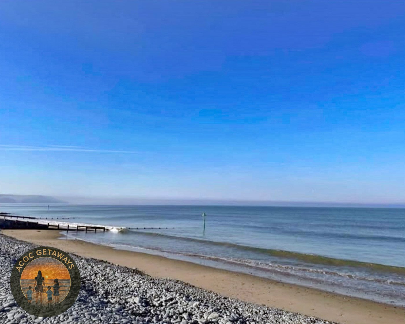 A peaceful beach scene with pebbles in the foreground, a sandy shoreline, a calm ocean, and a clear blue sky.