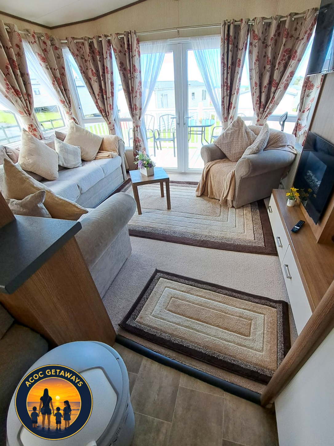 Living room with beige sofas, a wooden coffee table with flowers, patterned curtains, a TV on a wooden stand, and sliding glass doors leading to a balcony with outdoor chairs.