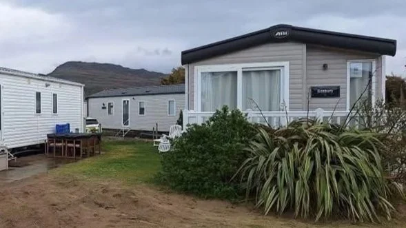 A campground with several mobile homes on grassy lots surrounded by bushes and plants, with a mountain in the background and cloudy sky.