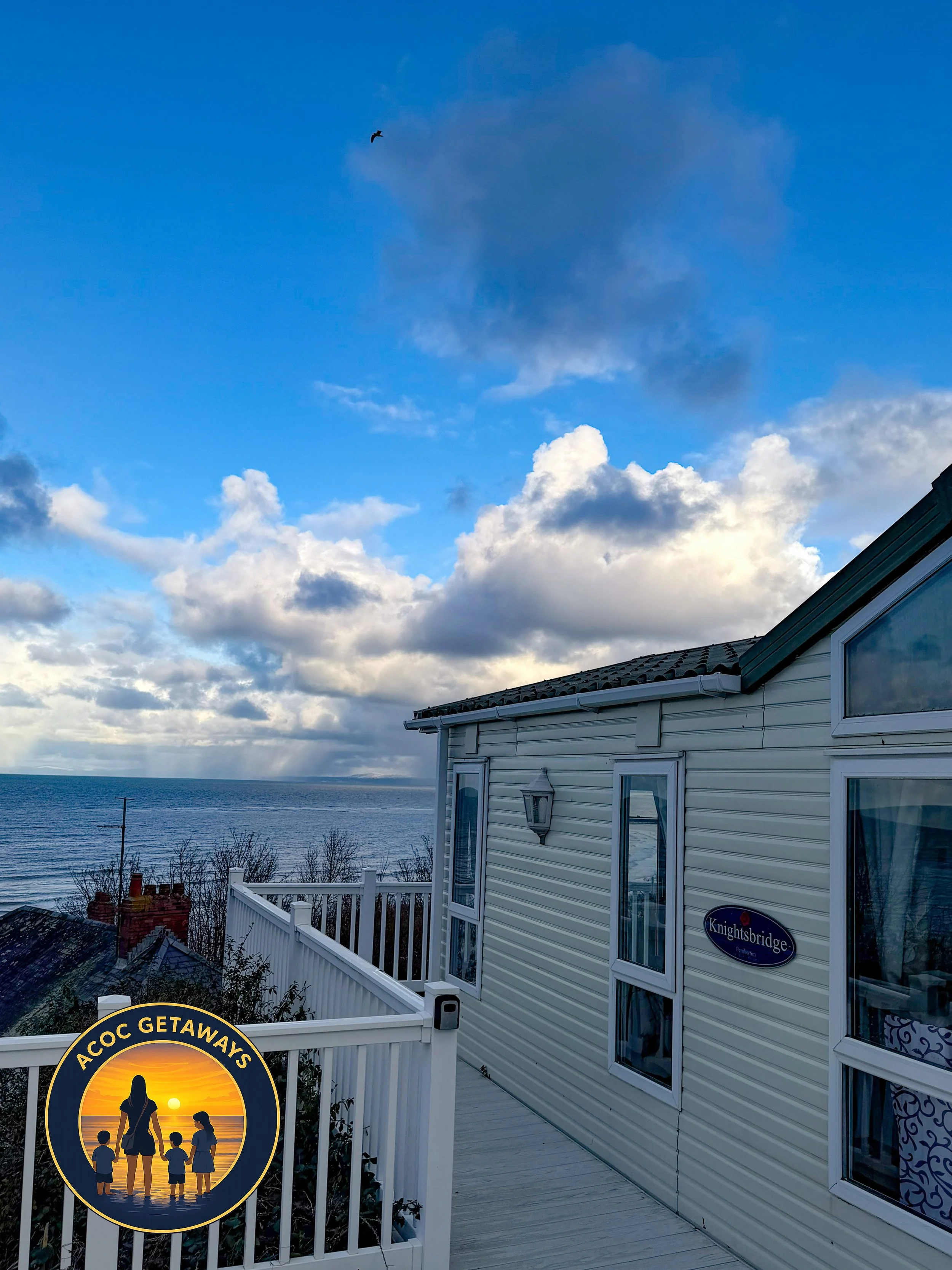 A balcony view overlooking the ocean with cloudy sky and a house. There is a sign on the fence that reads 'ACOC GETAWAYS' with a silhouette of a woman with children against a sunset background.