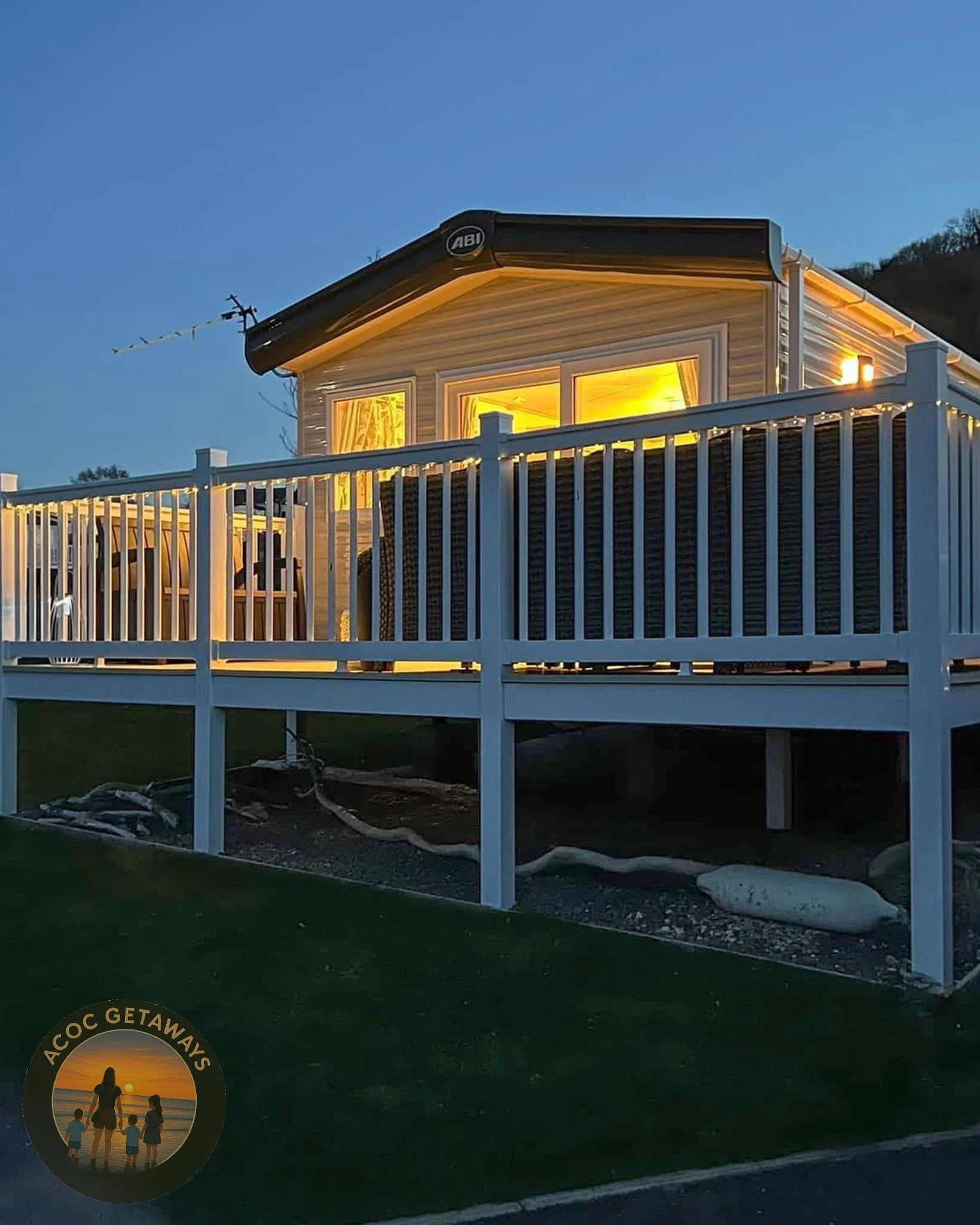 A house with an illuminated interior seen through windows, a white deck with outdoor furniture, a grassy yard, and a logo featuring a family at sunset with the text "Accoc Getaways".