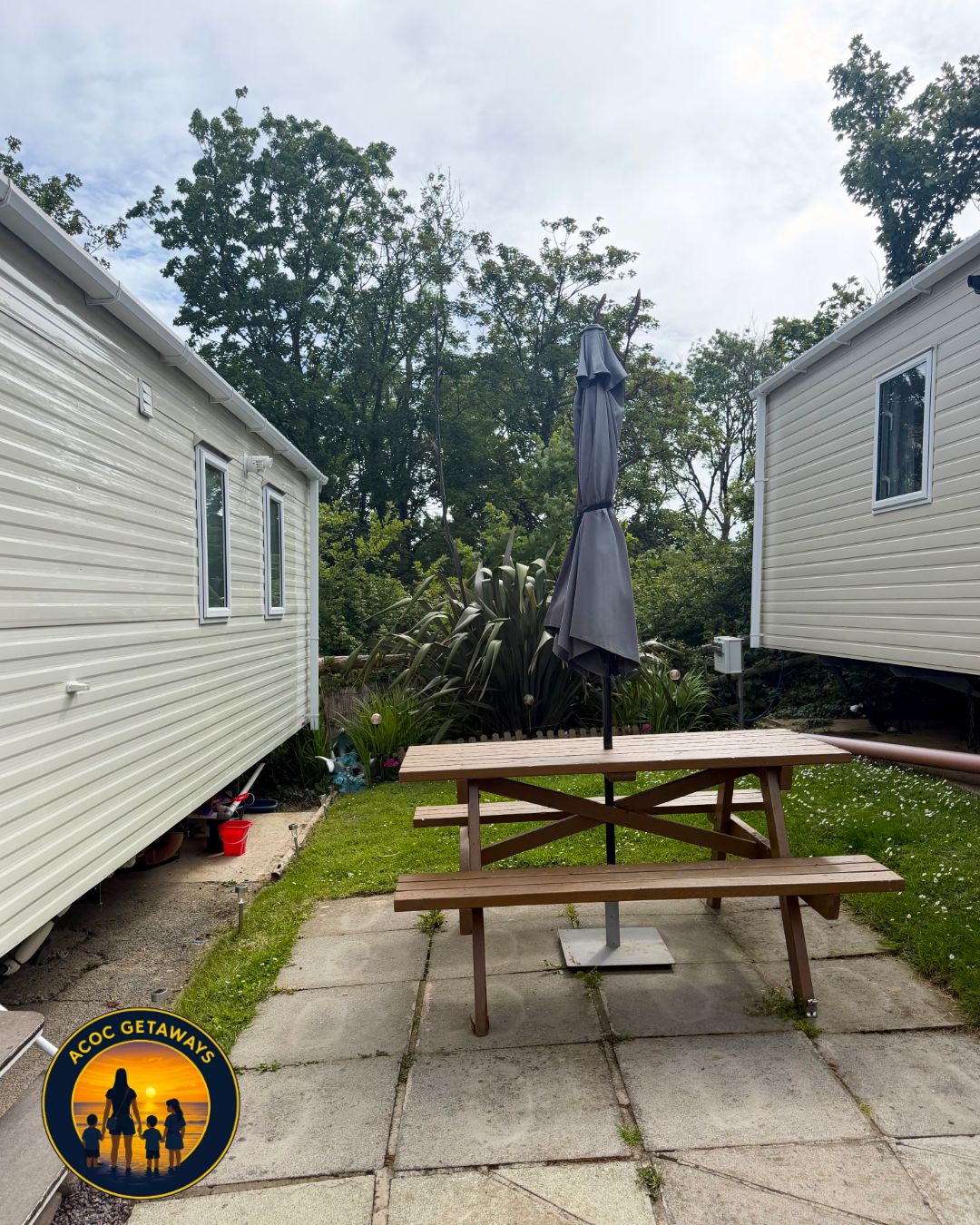 Backyard patio with wooden picnic table and closed umbrella, surrounded by mobile homes, greenery, and trees in the background.