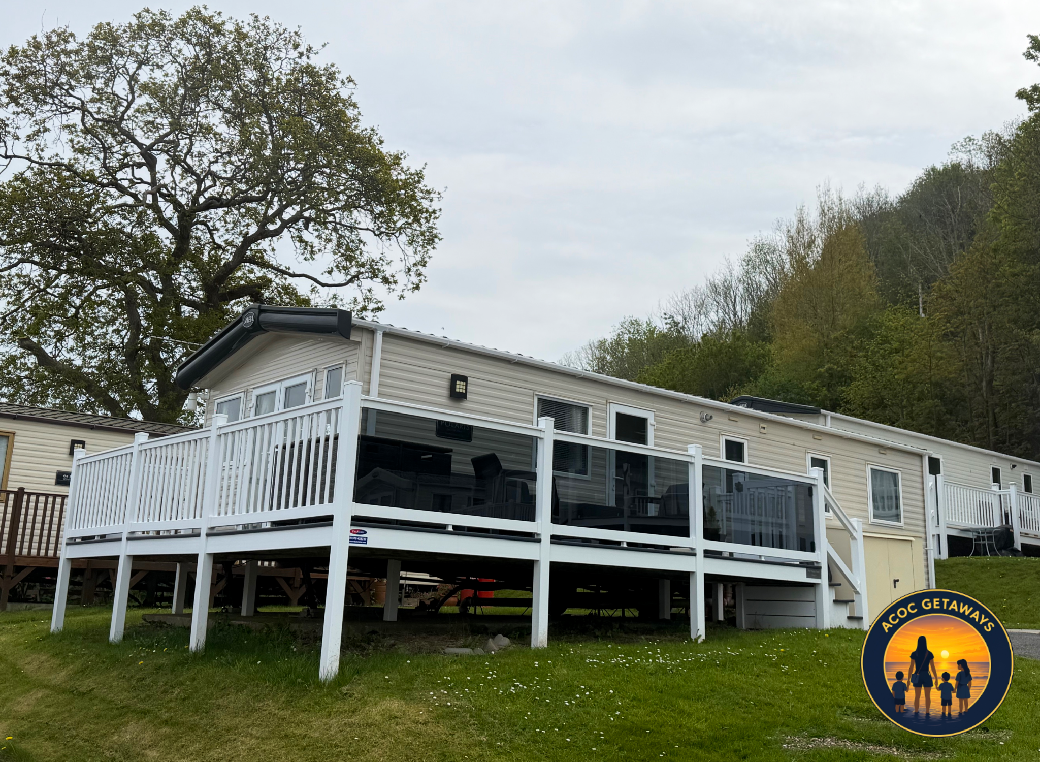 A residential house with white siding and a deck, surrounded by green grass and trees under a cloudy sky.