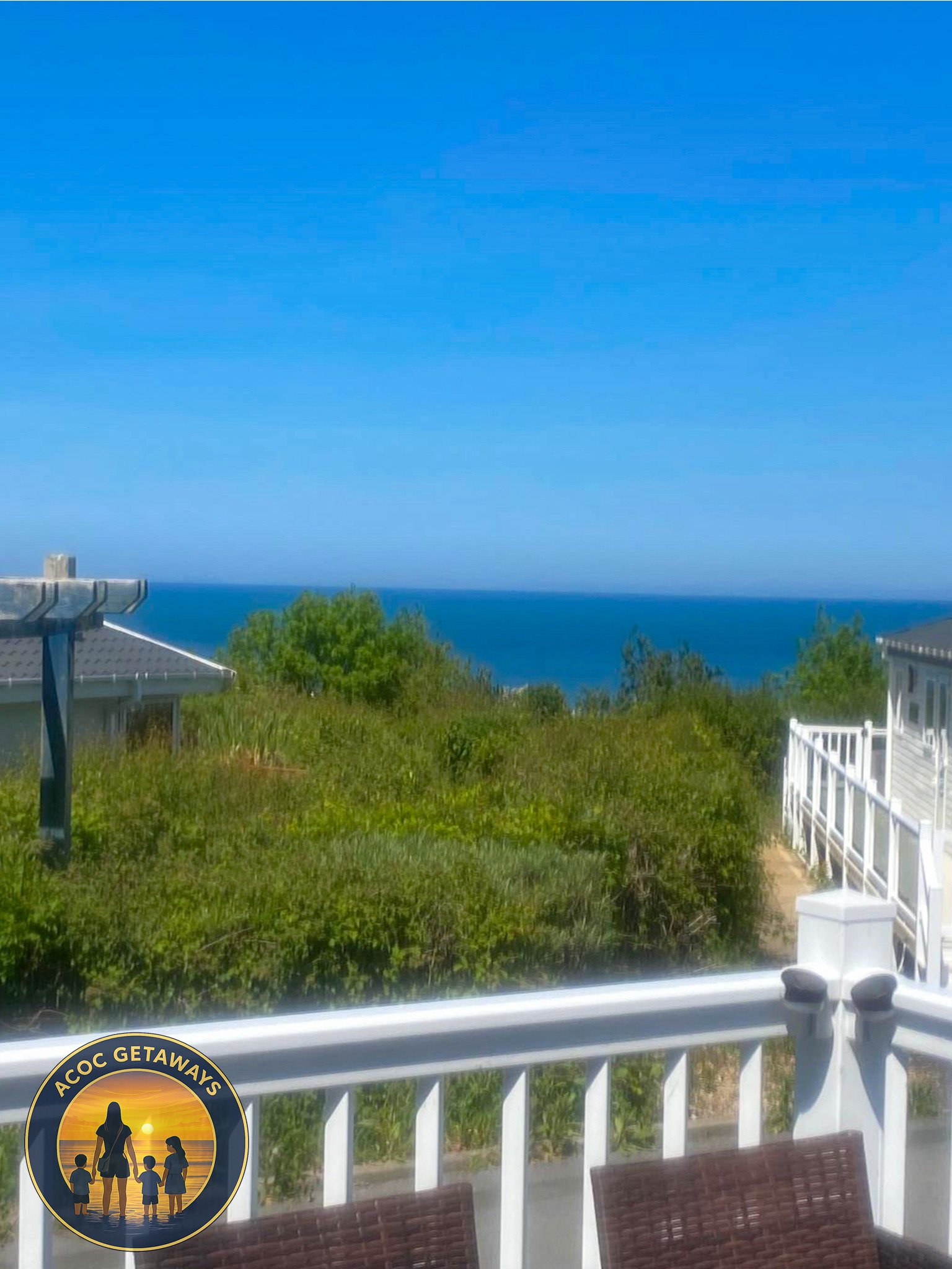 Ocean view from a balcony with white railing, green bushes, a house with a gray roof, and the sky in the background.