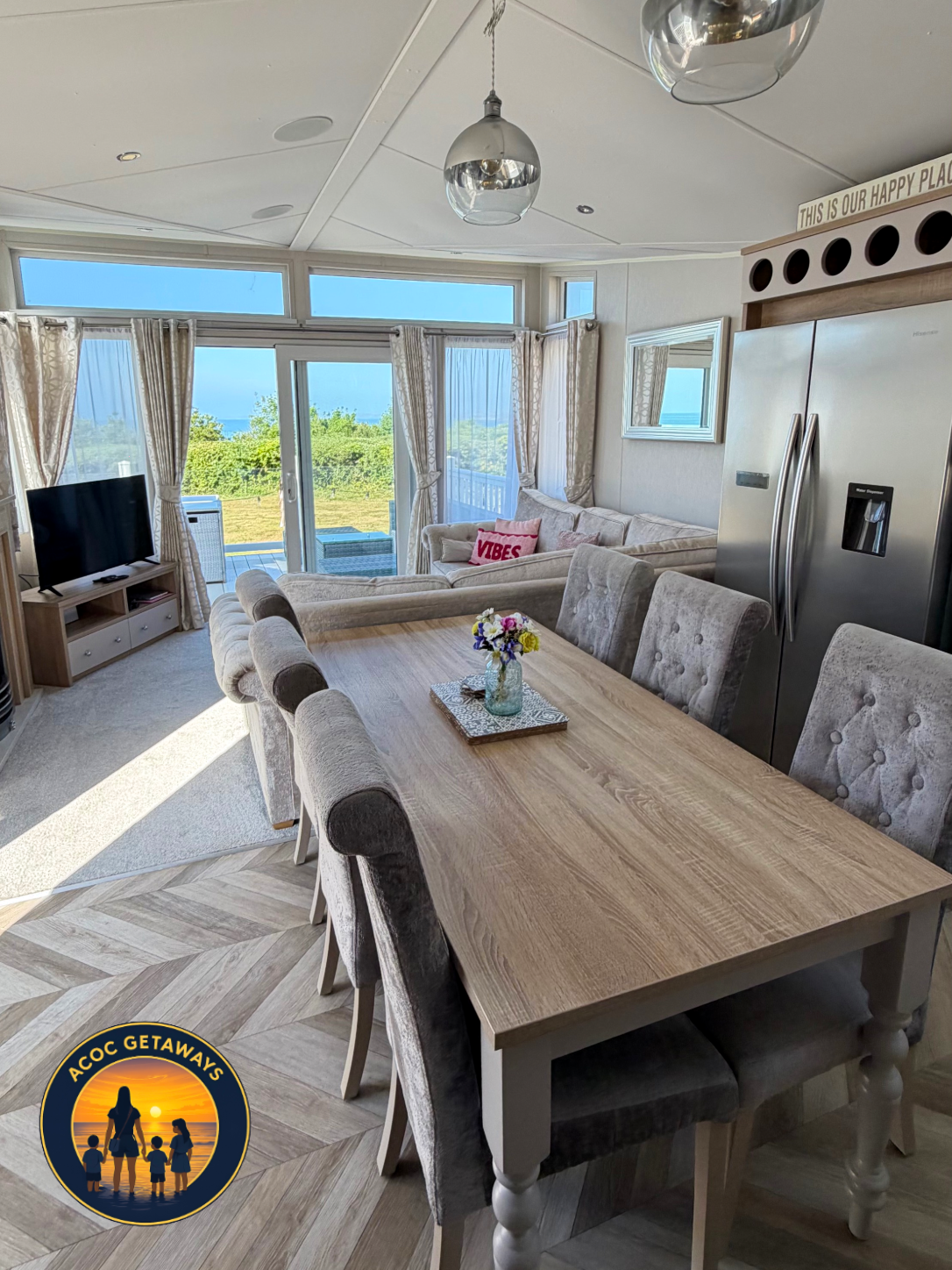 A bright living and dining room with a large wooden table, beige upholstered chairs, a sofa with pink and red pillows, a flat-screen TV, and ocean views through glass sliding doors and windows.