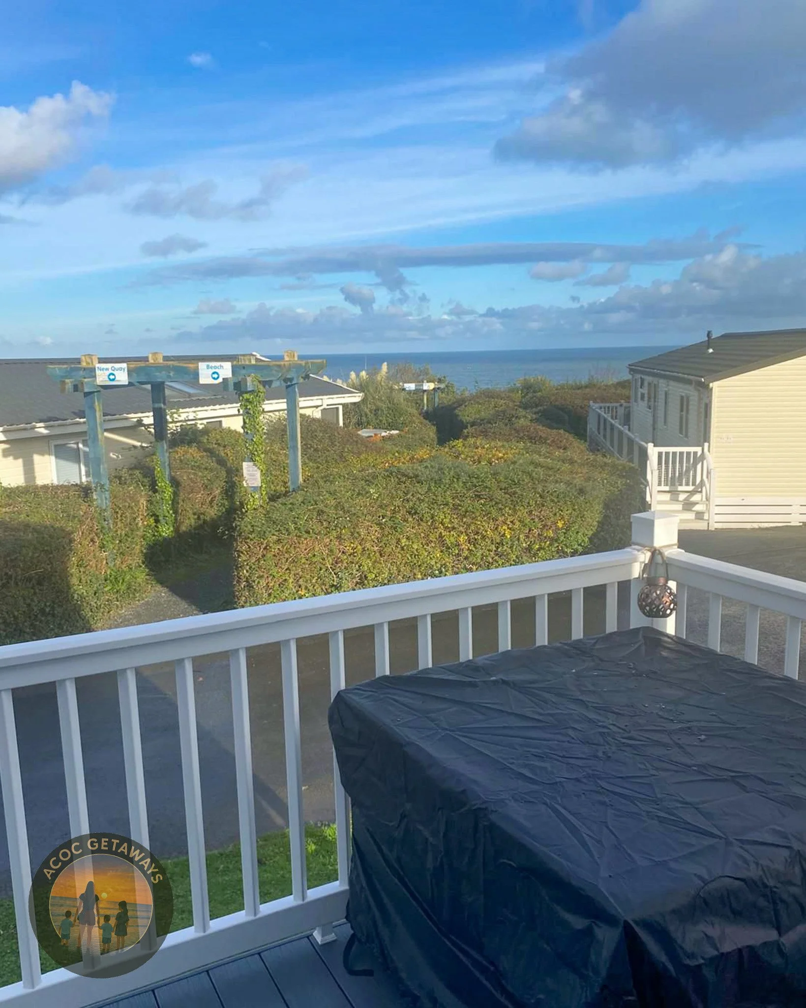 View from a balcony showing a black covered grill, white railing, bushes, a wooden sign structure with signs labeled 'New Quay' and 'Beach,' neighboring houses, and the ocean in the distance under a blue sky with scattered clouds.