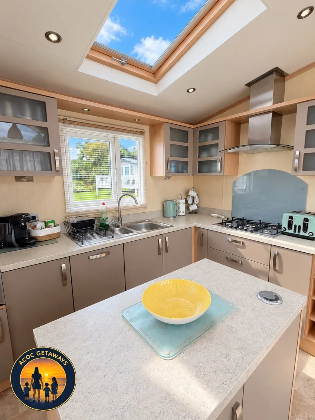 A bright kitchen with a skylight showing a blue sky with clouds, beige cabinets with frosted glass doors, a window with blinds, a countertop with small appliances and utensils, a yellow bowl on a cutting board, and a logo in the bottom left corner th