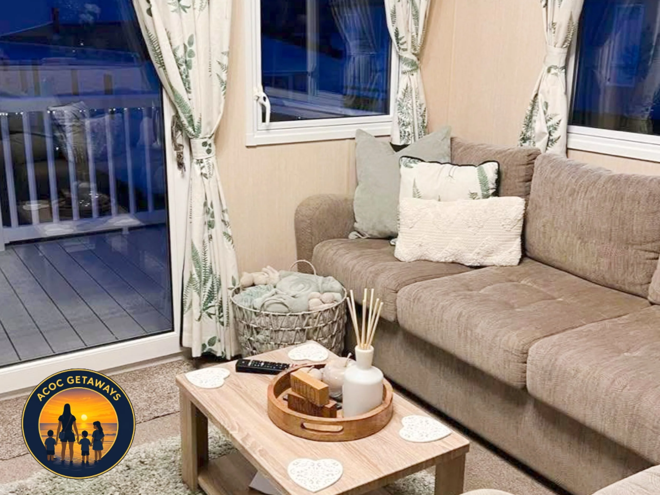 Living room with beige sectional sofa, pillows, a coffee table with a round wooden tray, and a basket of plush toys near a sliding glass door with floral curtains. Outside the door, a balcony with railing is visible.