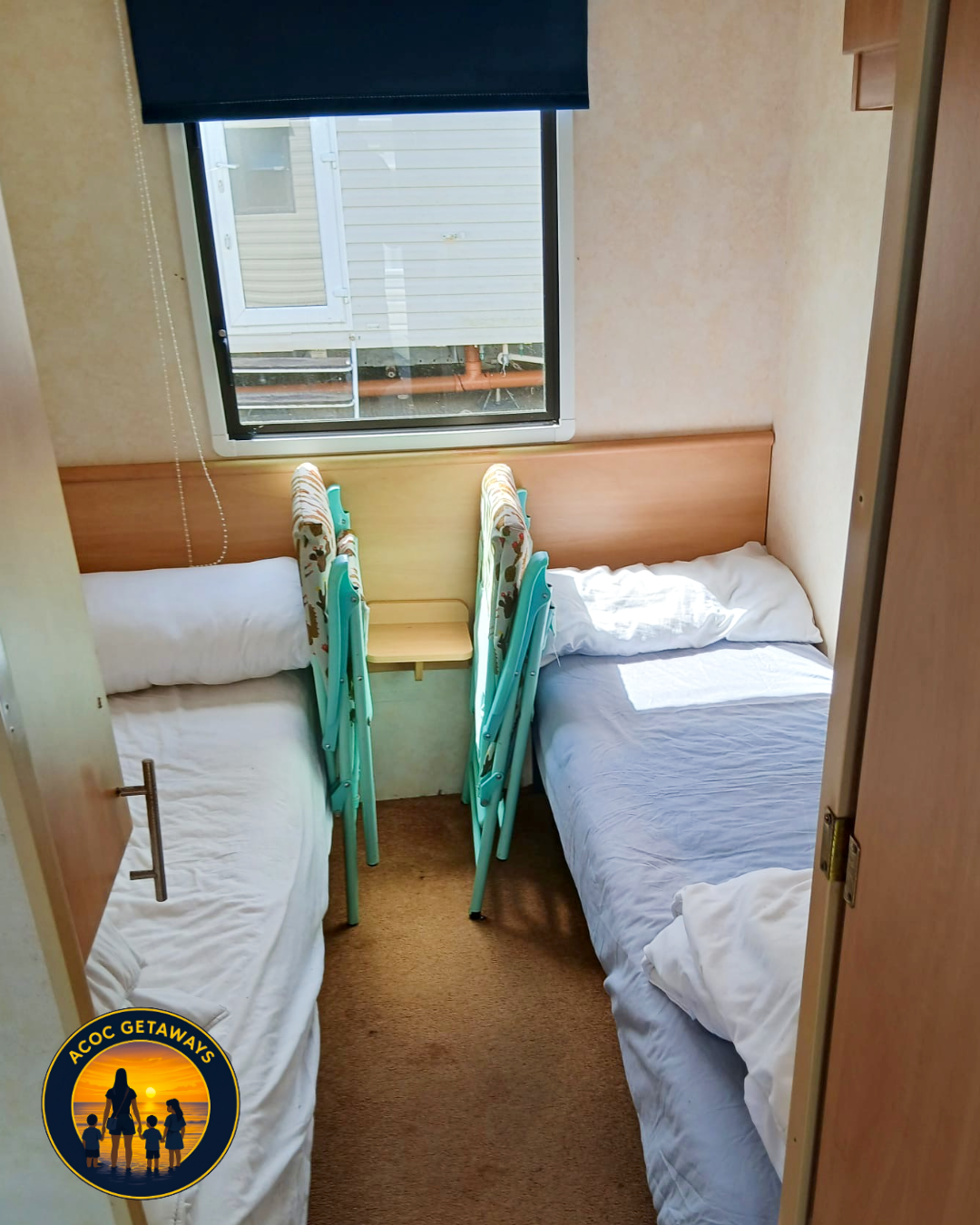 Small bedroom with two single beds, a window with a blue blind, and stacked chairs at the foot of the beds.