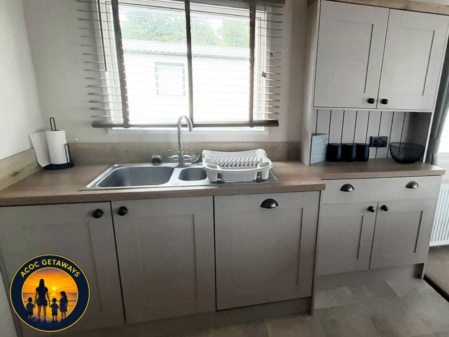 Kitchen with beige cabinets, a window above the sink, a paper towel holder, a dish drainer, and various black kitchen accessories.