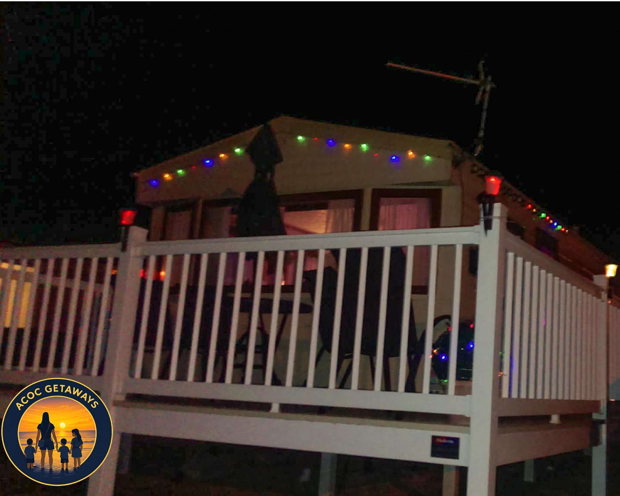 Nighttime view of a two-story house with a decorated balcony, string lights along the roofline, and a flagpole with an antenna. The lower level has a white railing, and there are chairs and a bicycle visible behind it. There is a logo in the bottom l