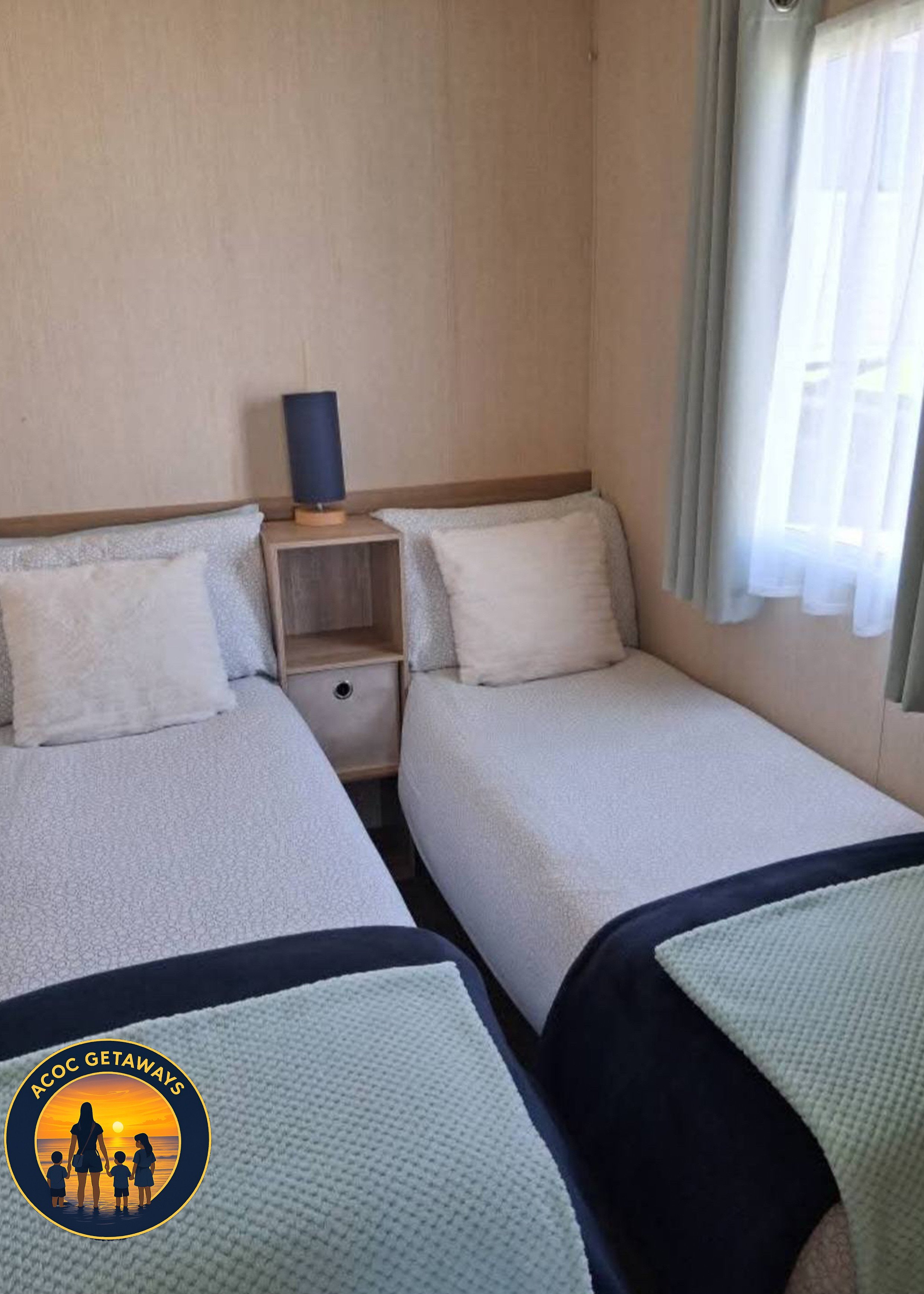 A room with two twin beds with beige bedding and pillows, a small wooden nightstand with a drawer, and a black table lamp. There is a window with sheer white curtains and light green drapes, and a round logo with a sunset and family silhouette is in 