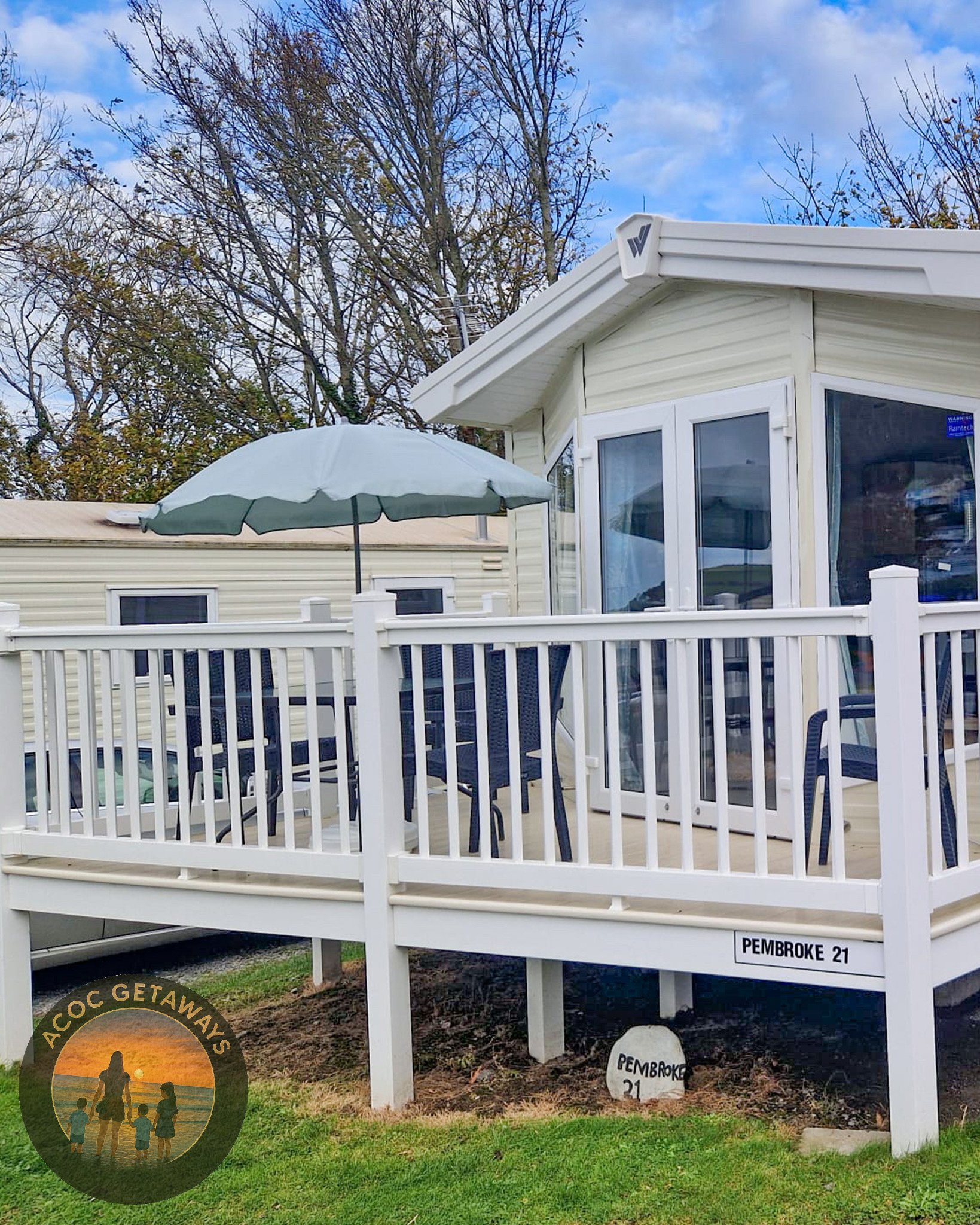 A manufactured home with a porch, black chairs, an umbrella, and a white fence, with a sign labeled 'PEMBROKE 21' and a round stone with the same label in the yard.