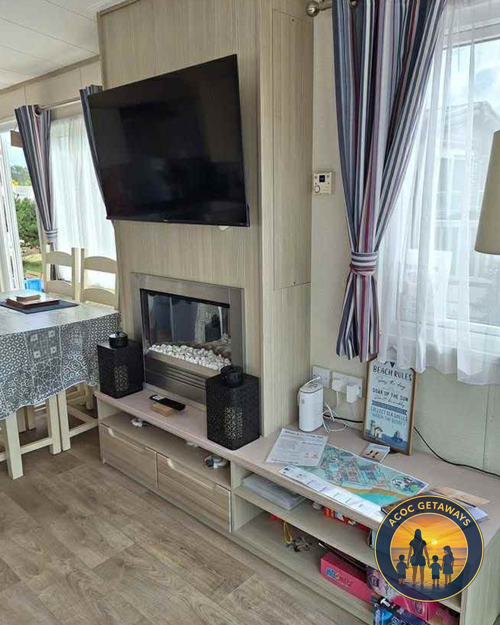 Living room corner with wall-mounted flat-screen TV, fireplace below, white wooden TV stand with storage, window with striped curtains, side table with magazines, and wall outlet with plugged-in devices.