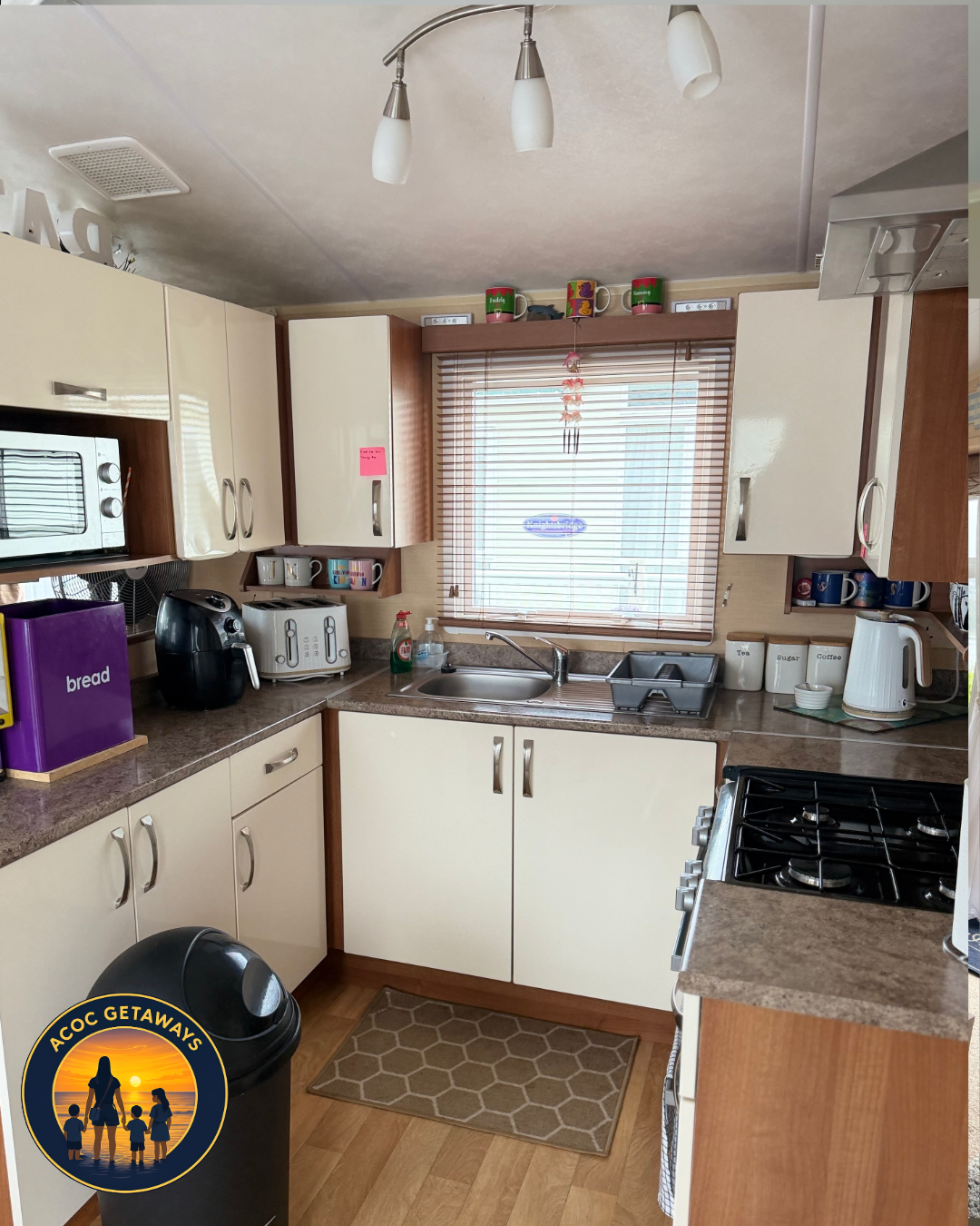 Kitchen with white cabinets, brown countertops, a window with blinds, and various appliances including a toaster, coffee maker, microwave, and kettle. There is a trash can and a beige rug on the wooden floor.