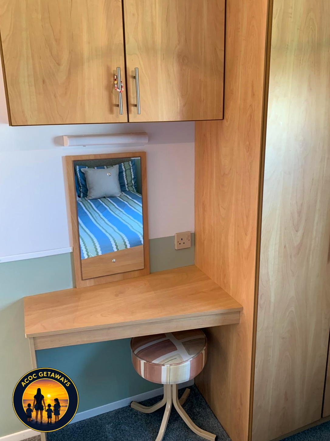 Wooden desk and cabinetry in a hotel room or accommodation, with a small mirror reflecting a bed with green striped bedding and a gray pillow, and an upholstered stool.