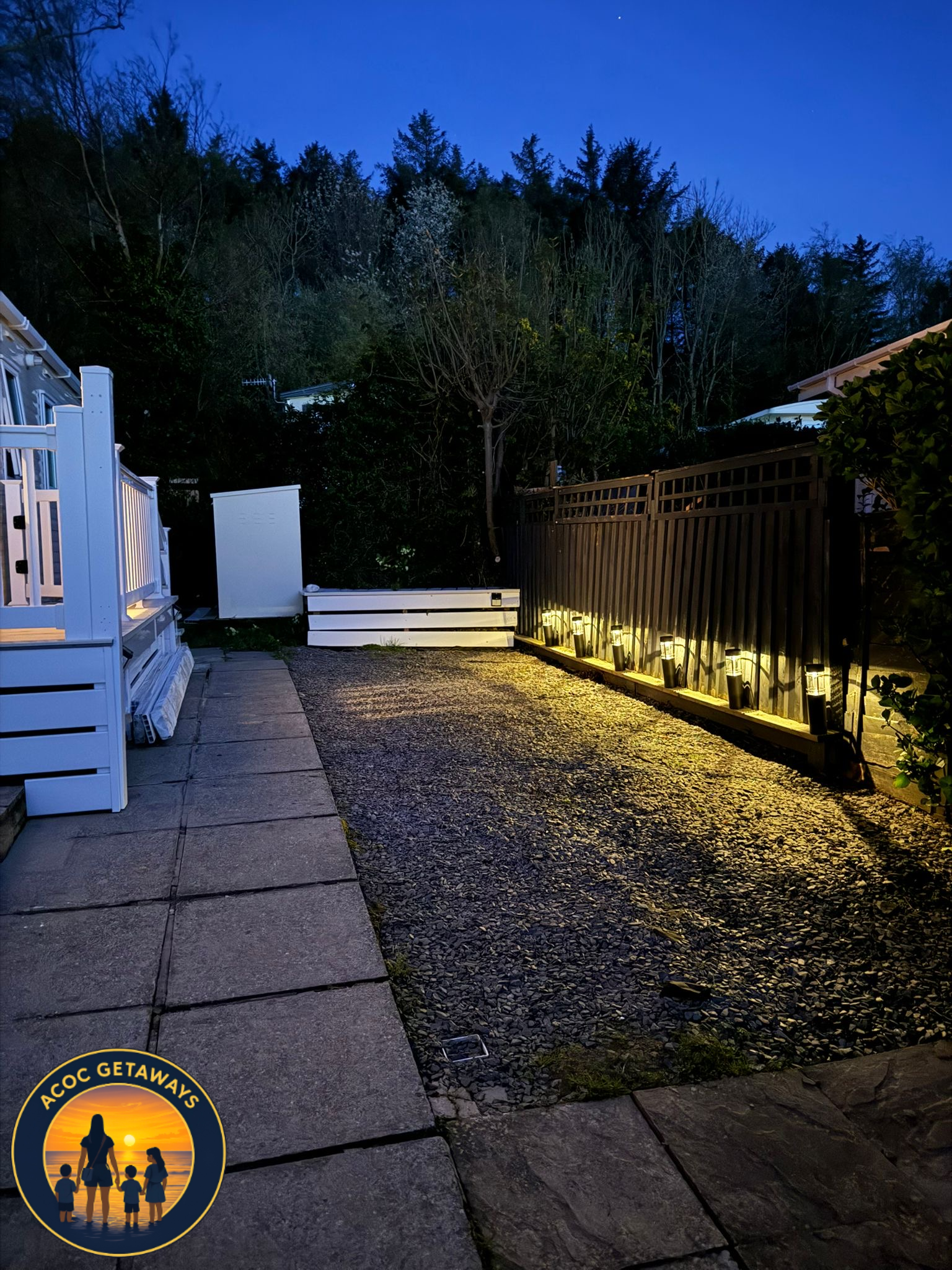 A backyard patio area at dusk with outdoor lighting along the black fence, a tree and wooded area in the background, and a combination of stone and gravel ground surface.
