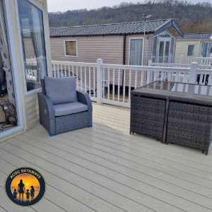 Photo of a balcony or deck with a gray armchair and a black wicker outdoor furniture set, enclosed with a white railing, overlooking neighboring houses and a hilly background.