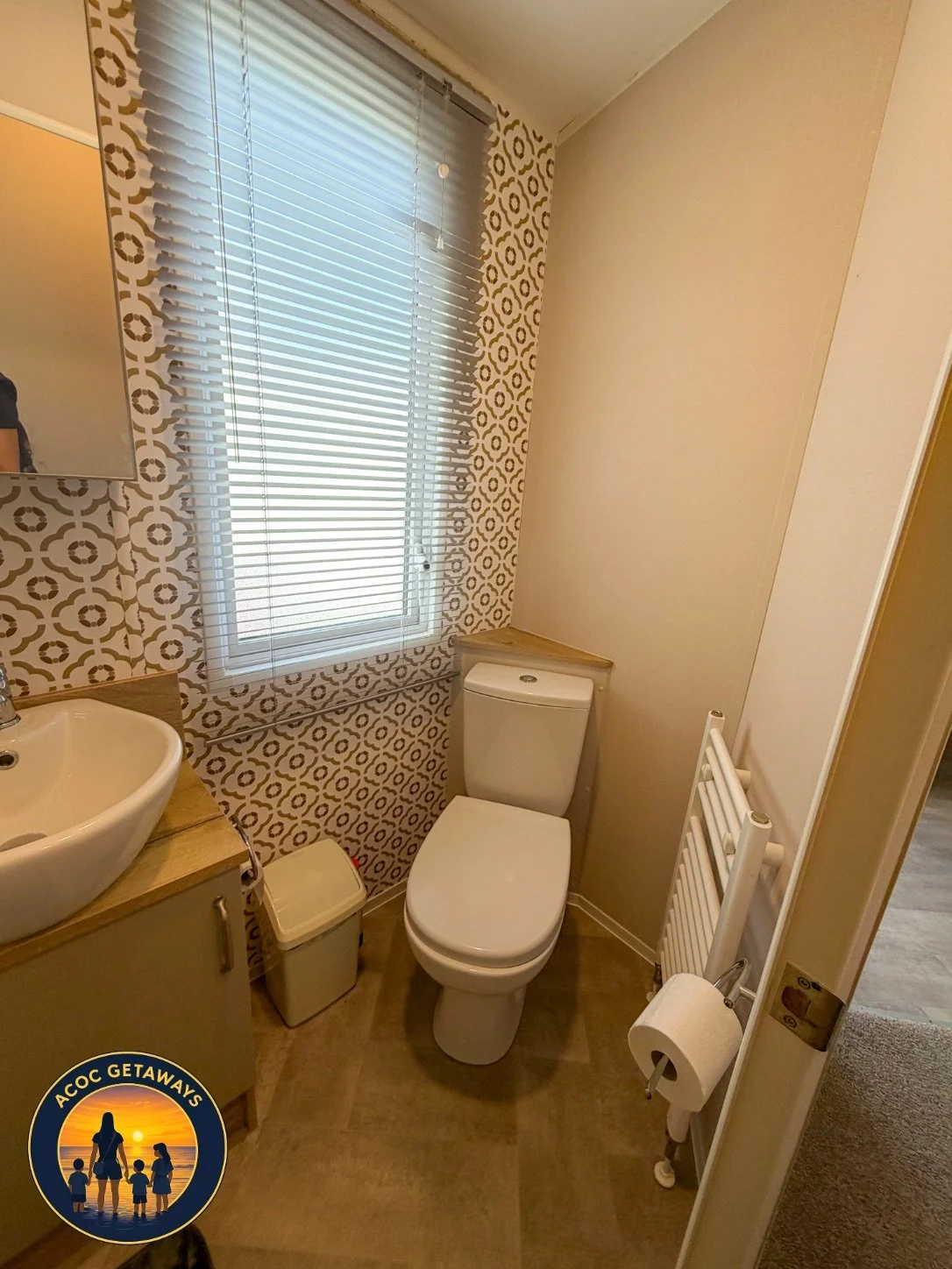 A small bathroom with a window, patterned wall tiles, a white toilet, a small sink with a wooden cabinet, a trash bin, and a white radiator with a toilet paper roll, seen from the doorway.