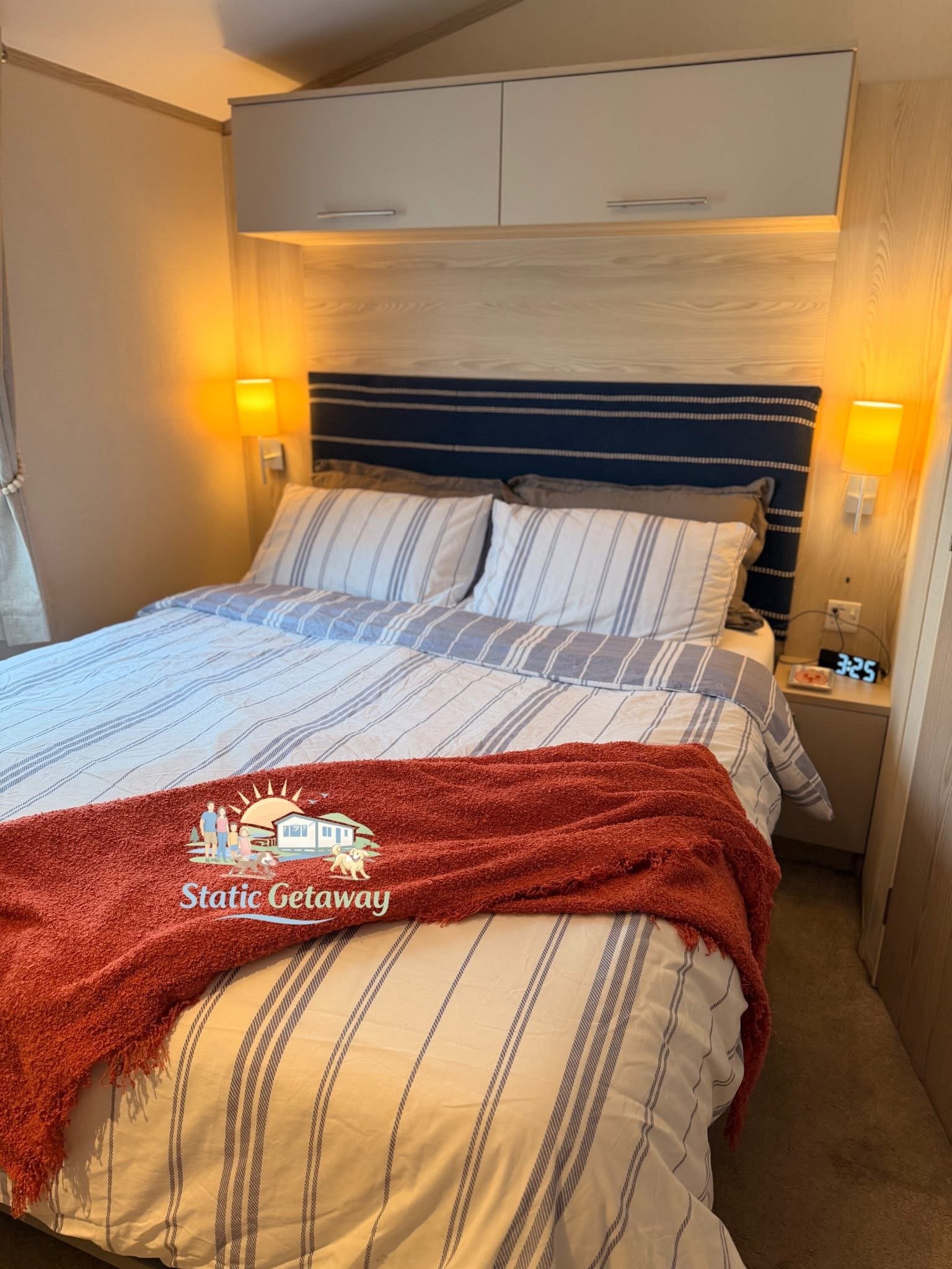 A neatly made bed with a striped sheet, brown pillows, and a red throw blanket. There are two wall-mounted yellow lamps on either side of a headboard, which is padded and covered with striped fabric. A small bedside table with a digital clock and a f