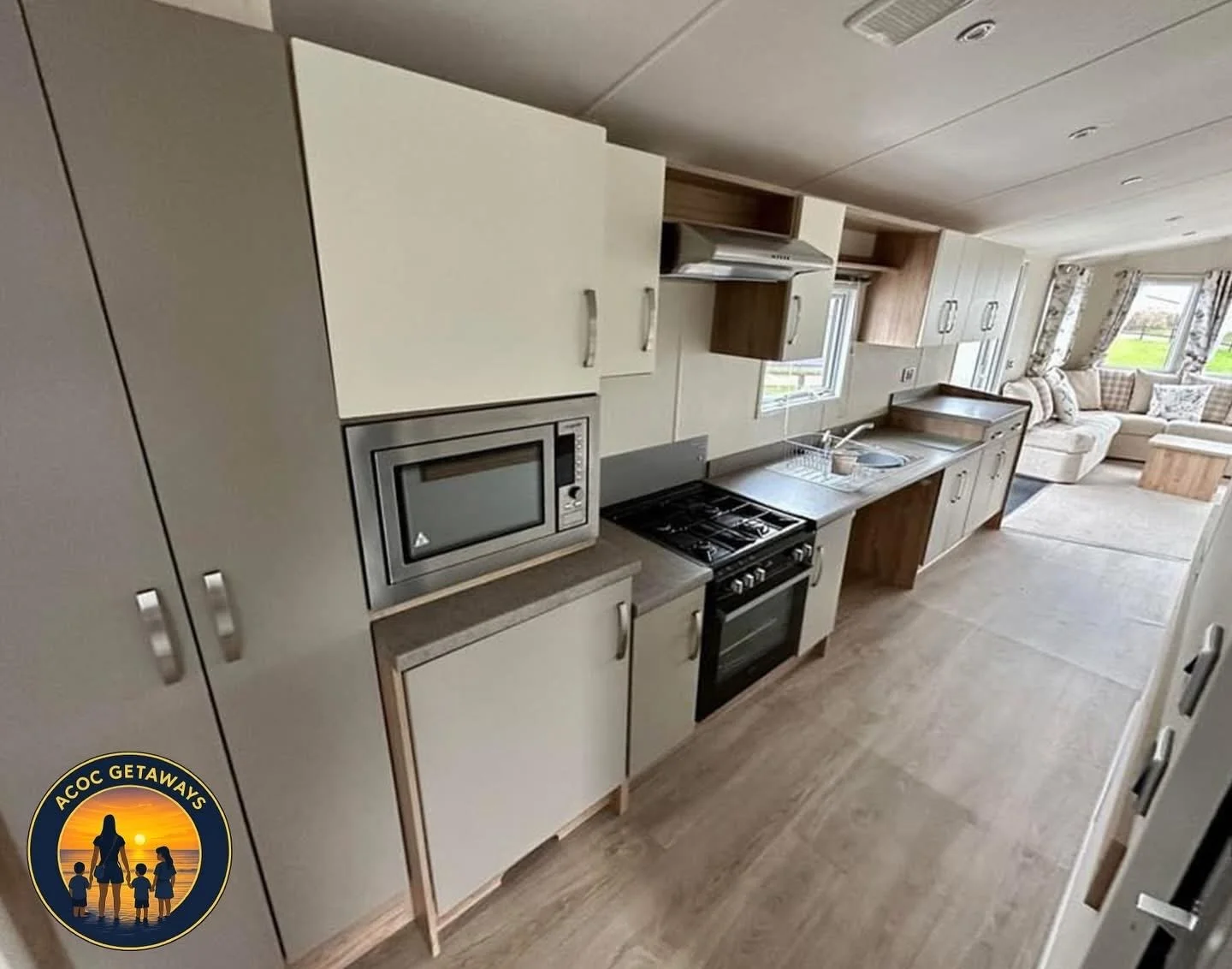 A cozy open-concept kitchen with white and wooden cabinets, a microwave, a stove, a sink, and a view of a living area with a sectional sofa and large windows showing a grassy outdoor area.