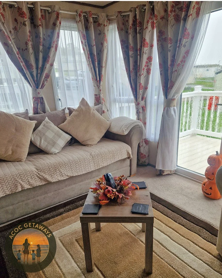 Living room with beige sofa, pillows, lace curtains with floral patterns, sliding glass door with outdoor view, pumpkin decorations on the right, small wooden coffee table with autumn-themed centerpiece, and ACC COG GETAWAYS logo in the bottom left corner.