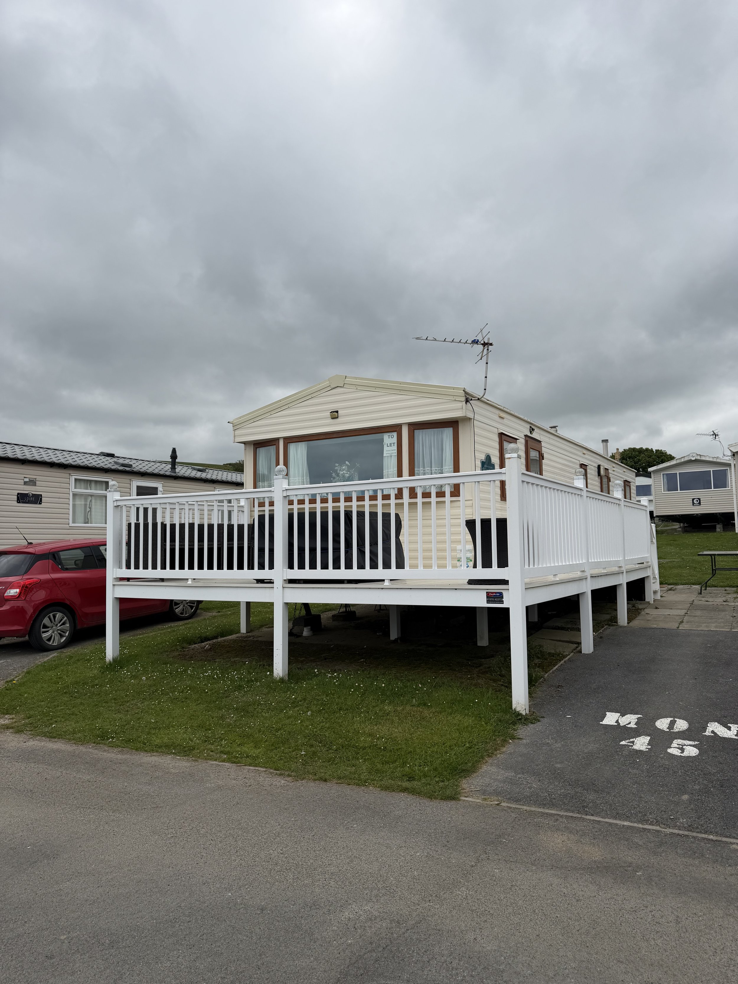 A beige mobile home with white railing on a cloudy day, parked on a lot with a 'TO LET' sign in the window, and a TV antenna on the roof. There are neighboring mobile homes and a red car nearby, with a parking space marked 'NO 45' in the foreground.