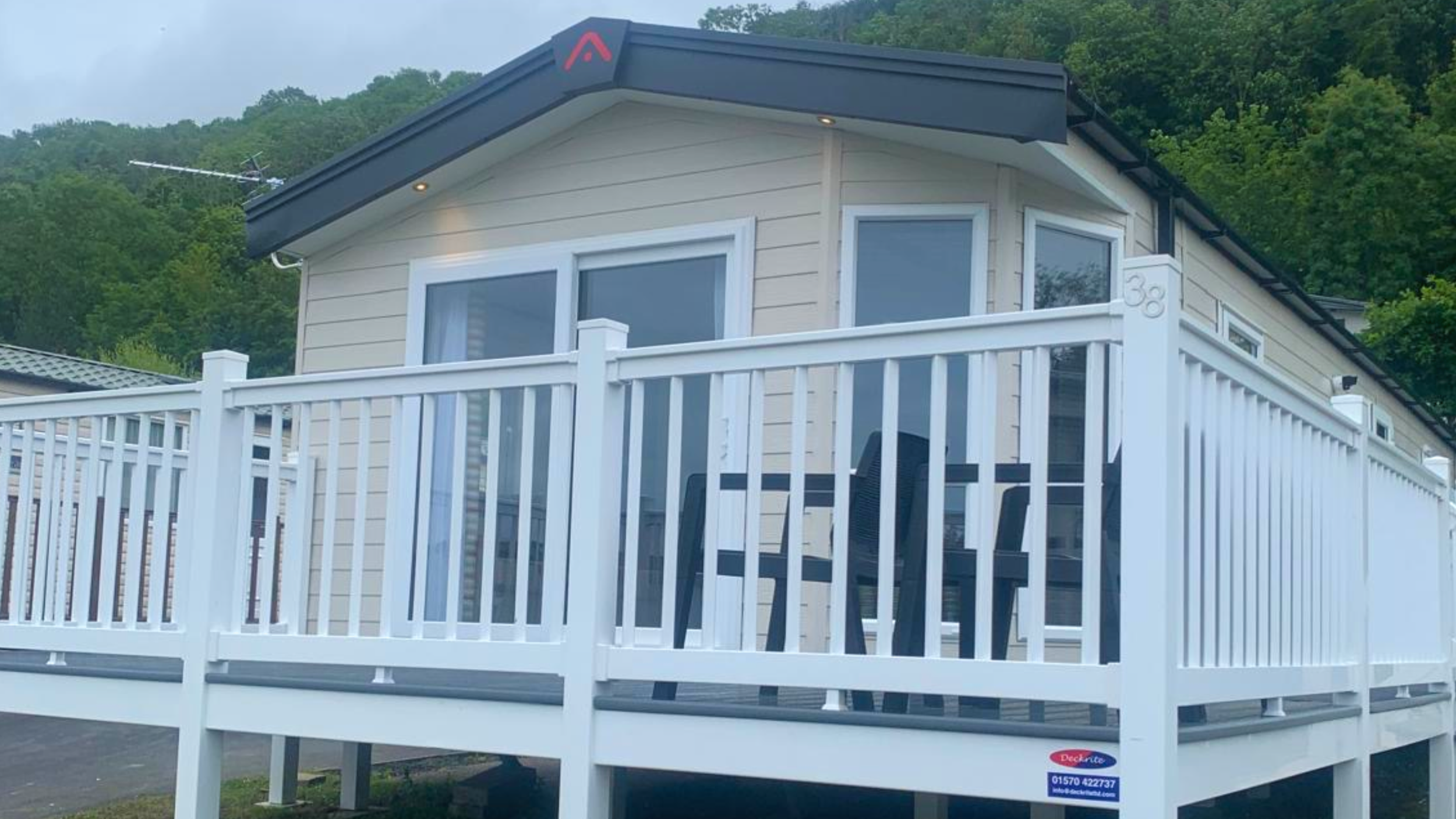 A beige house with a dark roof and white trim, featuring a balcony with white railing. The house is surrounded by green trees and hills in the background. There is a blue and red sticker on the bottom right corner of the house.