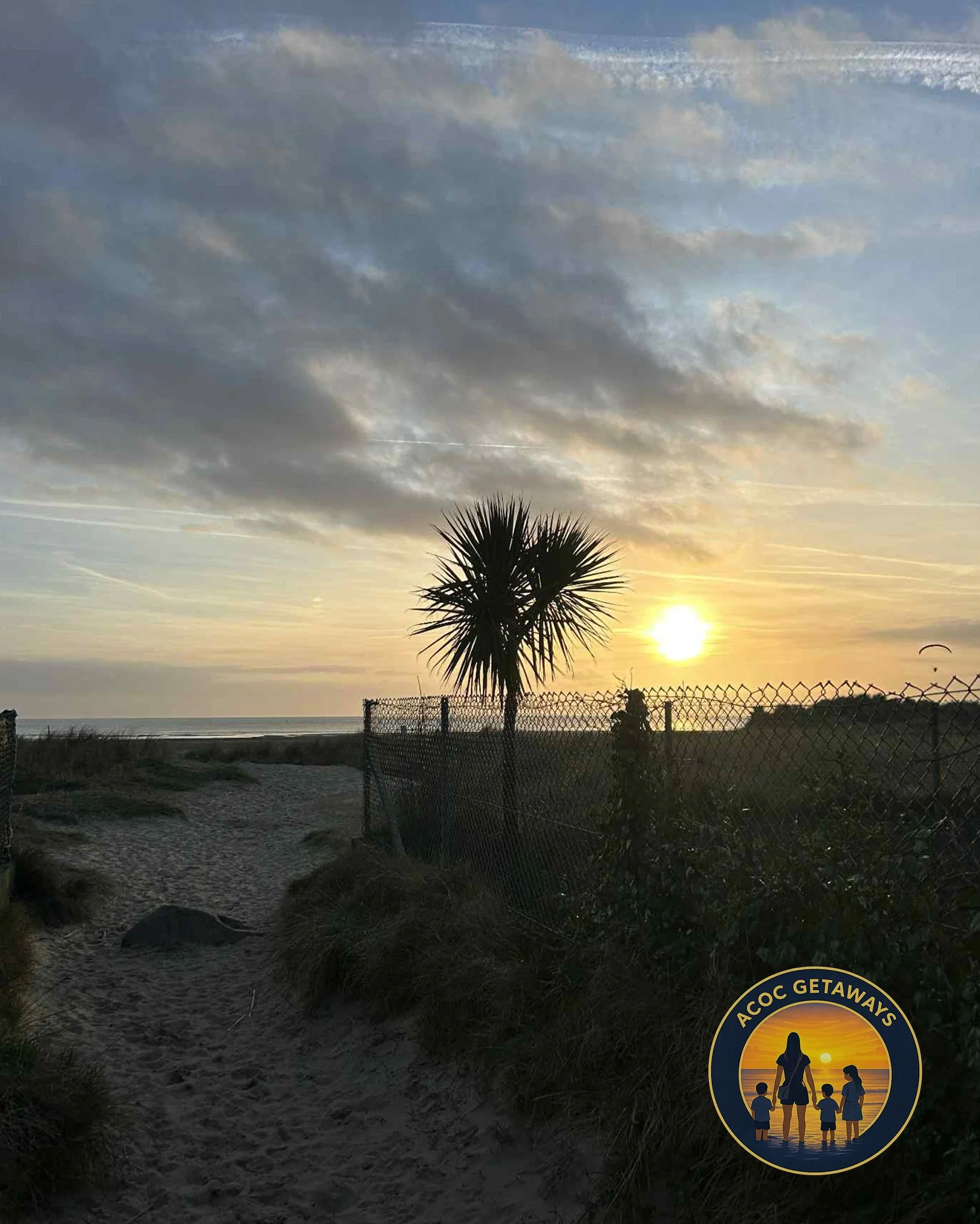 A sunset over a beach with a sandy path, a palm tree, a chain-link fence, grassy dunes, and a sky with clouds and contrails. There is a logo with a family silhouetted by the sunset in the bottom right corner.