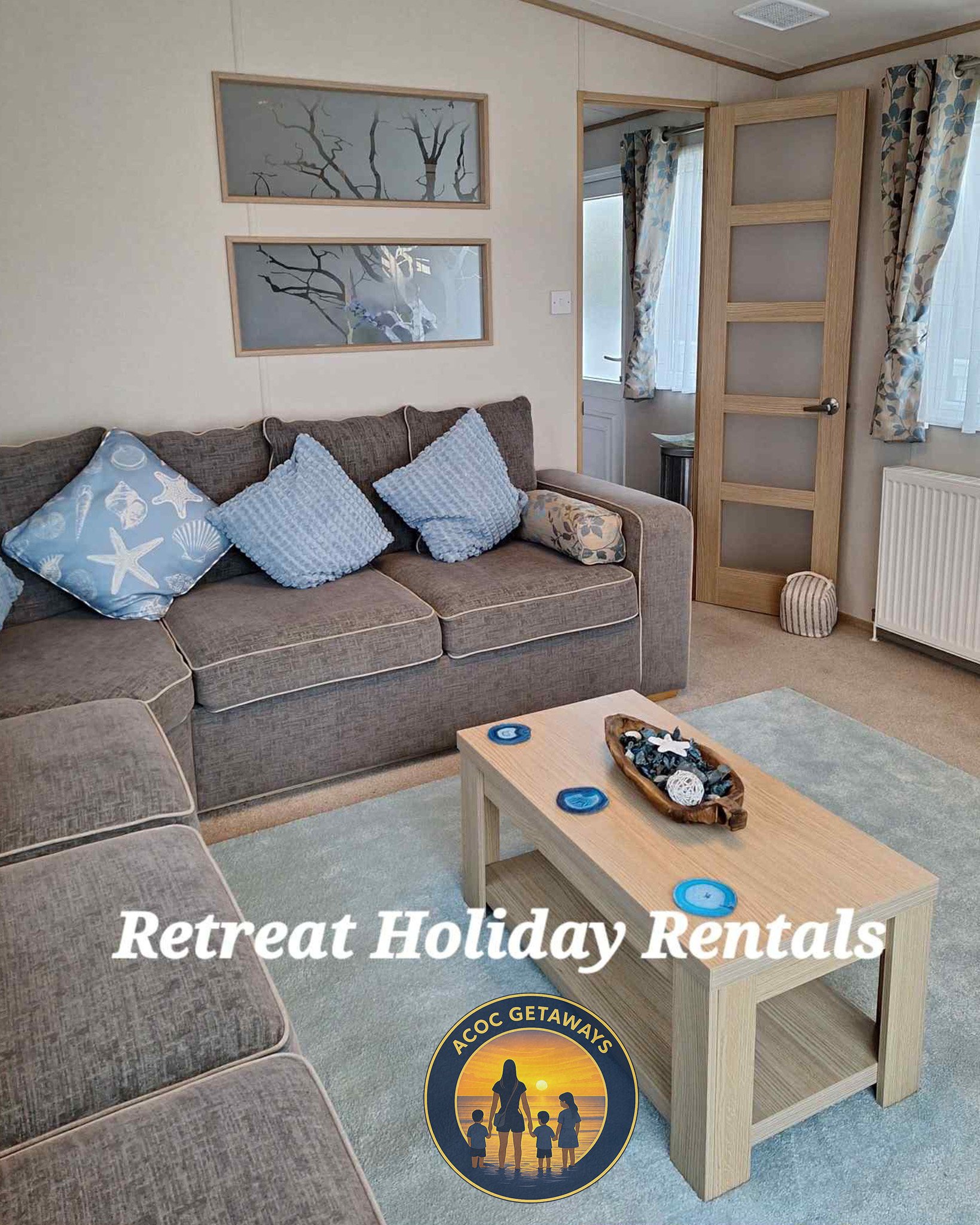 Living room with a gray sofa, blue pillows, a wooden coffee table with decorations, and a door leading to another room with curtains. The room has a light-colored rug and wall art featuring tree branches. Text overlay reads "Retreat Holiday Rentals" with an ACOV Getaways logo.