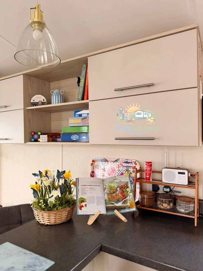 Kitchen countertop with a basket of daisies and yellow flowers, open recipe book on a stand, and a small wooden shelf with a radio, red container, and glass jars.