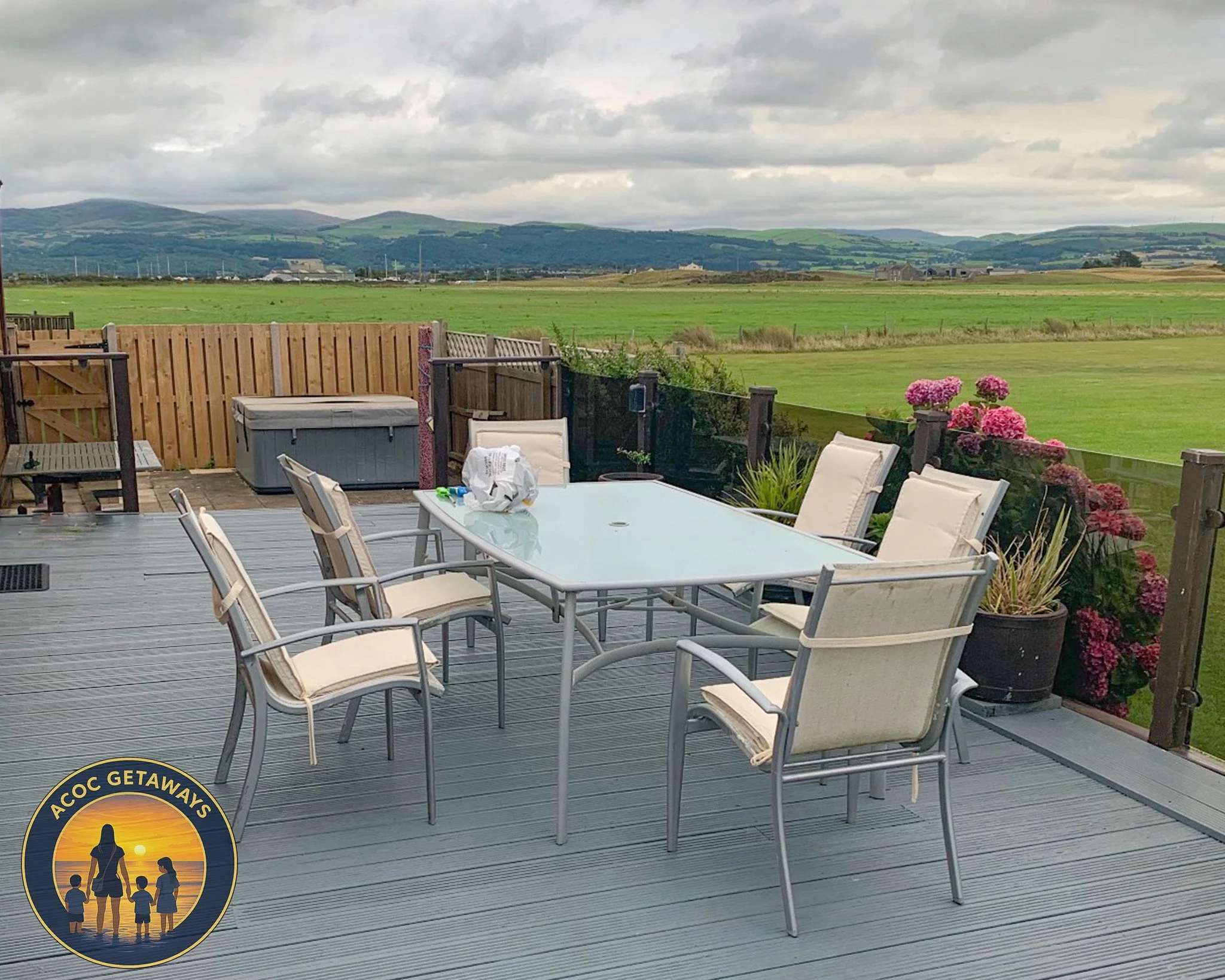 A backyard deck with a glass patio table, six chairs, pink hydrangea flowers in pots, and a green landscape with hills in the background under cloudy skies.