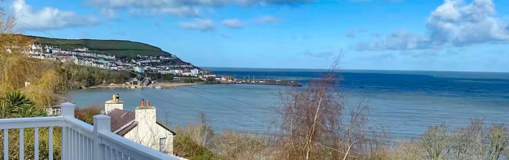 Scenic view of a coastal town with houses on a hillside overlooking the ocean, blue sky with scattered clouds, and a body of water with gentle waves.
