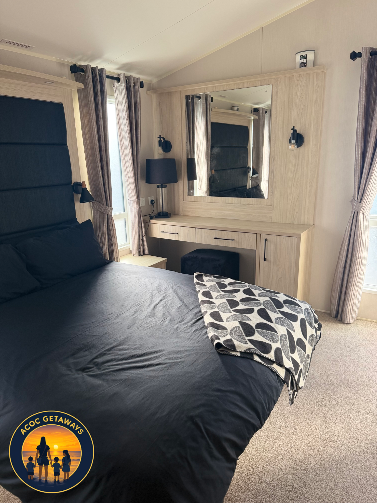 A bedroom with a bed covered in black bedding, a mirror, a nightstand with a black lamp, and curtains by the window