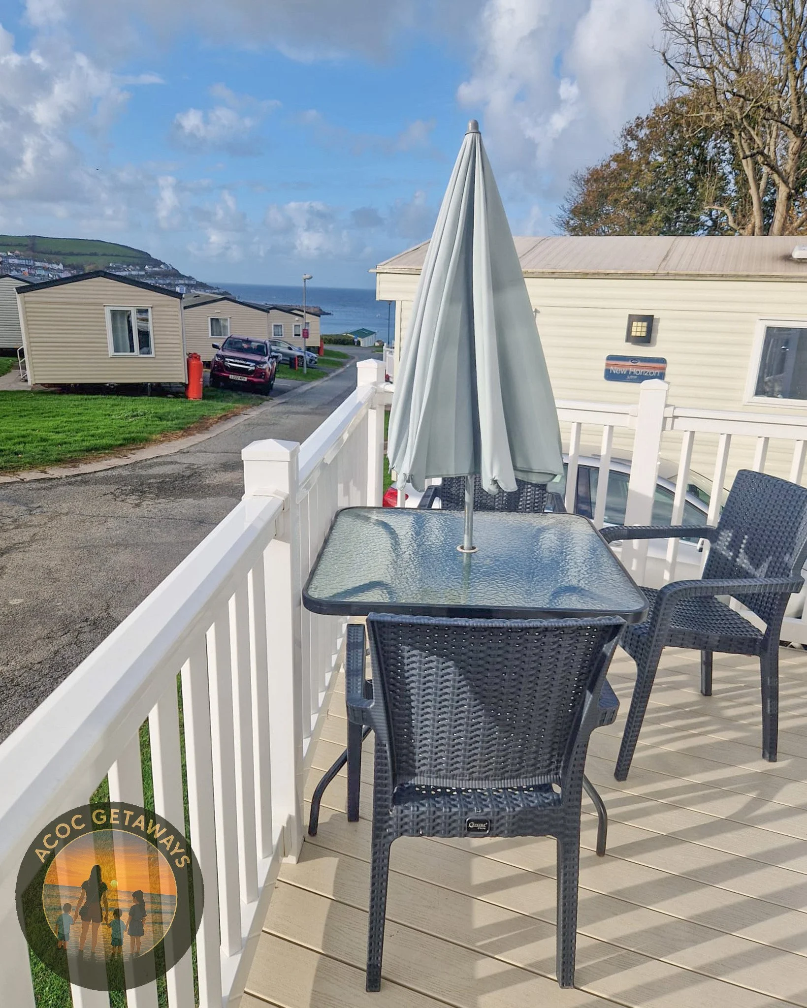 Outdoor balcony with a glass-top table, three wicker chairs, a large gray umbrella, and a view of neighboring houses, a hill, and the ocean in the background.