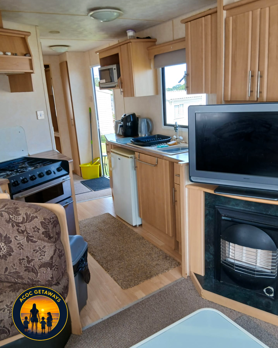 Interior view of a small kitchen in a recreational vehicle with wooden cabinetry, a stove, a mini-fridge, a sink, a microwave, and a television. There is a window above the sink, a door leading outside, a mop with a yellow bucket nearby, and various 