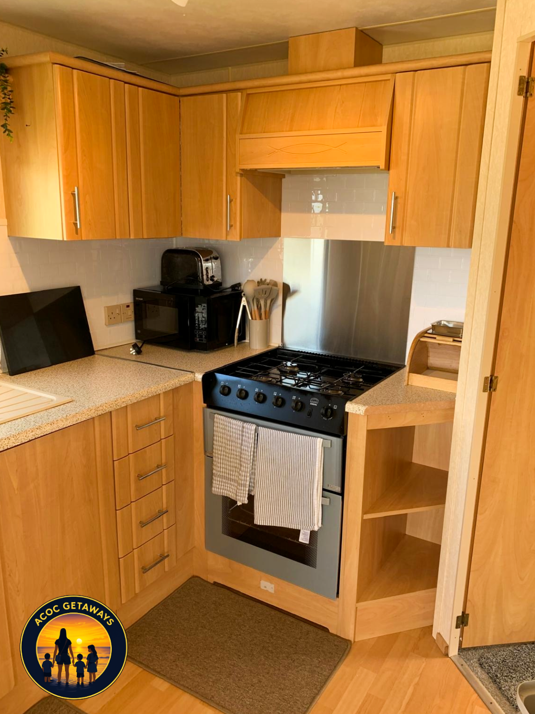 Kitchen with wooden cabinets, a stove with two towels hanging, a countertop with a dish drying rack, a black television, a microwave, a toaster, a utensil holder, and a small open corner shelf.