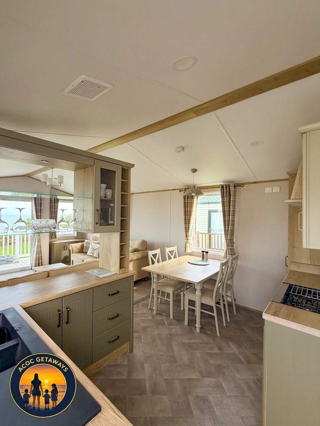 Open-concept kitchen and dining area in a modern home with beige and wood tones, a dining table with chairs, and large windows with plaid curtains.