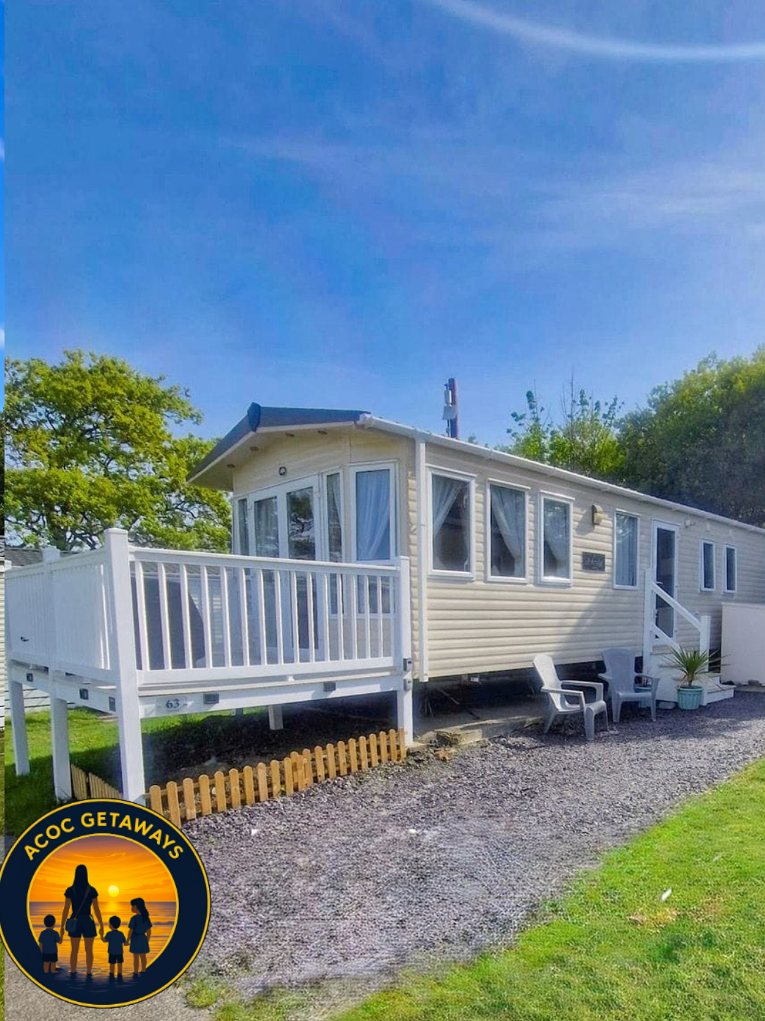 A white manufactured home with a small porch, outdoor seating, a potted plant, and surrounded by grass and trees under a bright blue sky.