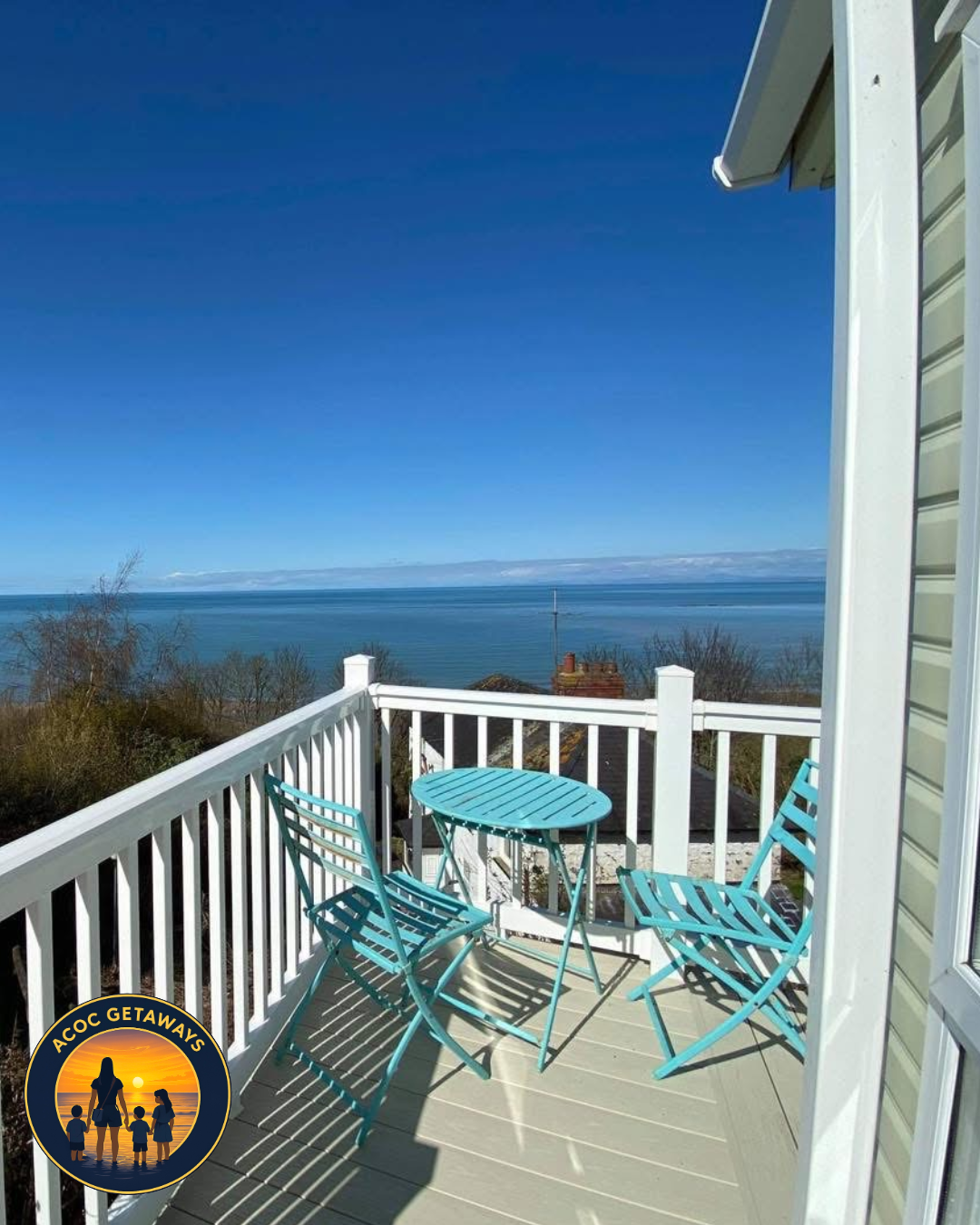 Balcony with a turquoise metal table and two chairs overlooking the ocean on a clear, sunny day.