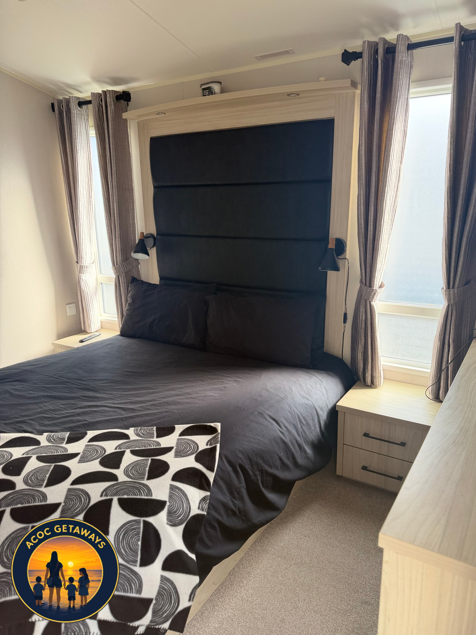 A bedroom with a bed that has black bedding, black pillows, and a black headboard. There are two nightstands with a lamp on each side of the bed. A window with beige and brown patterned curtains is behind the bed, and a second window with similar cur