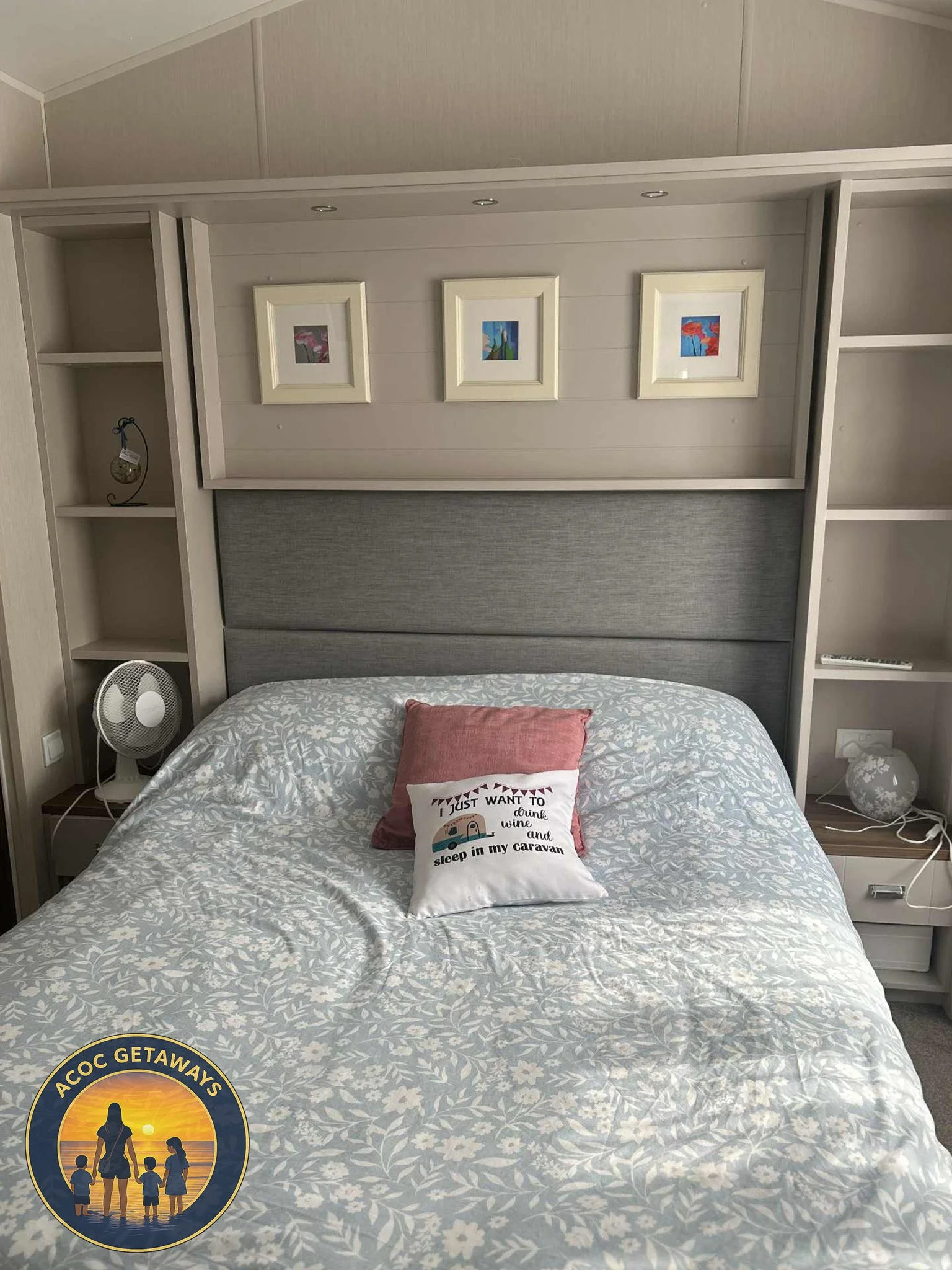 A neatly made bed with a patterned duvet cover, two decorative pillows, and a headboard in a cozy bedroom. Above the bed are three framed abstract art pieces, and there are built-in shelves and small nightstands on each side of the bed.