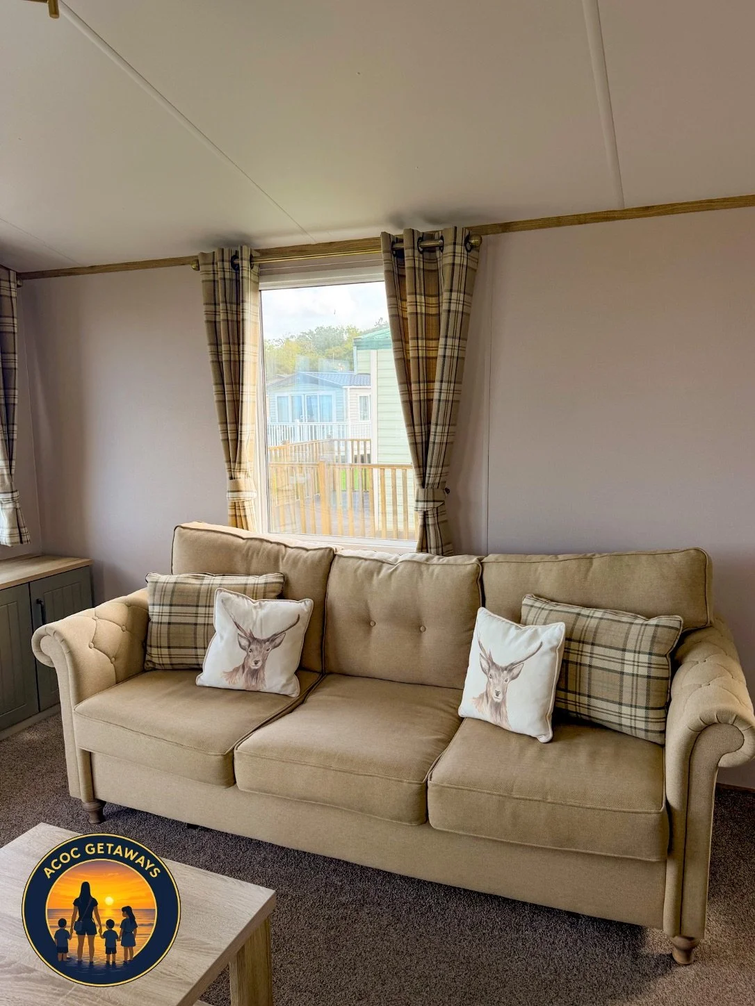 Living room with beige sofa decorated with deer-themed and plaid pillows, window with plaid curtains, view of a porch and other similar houses outside.