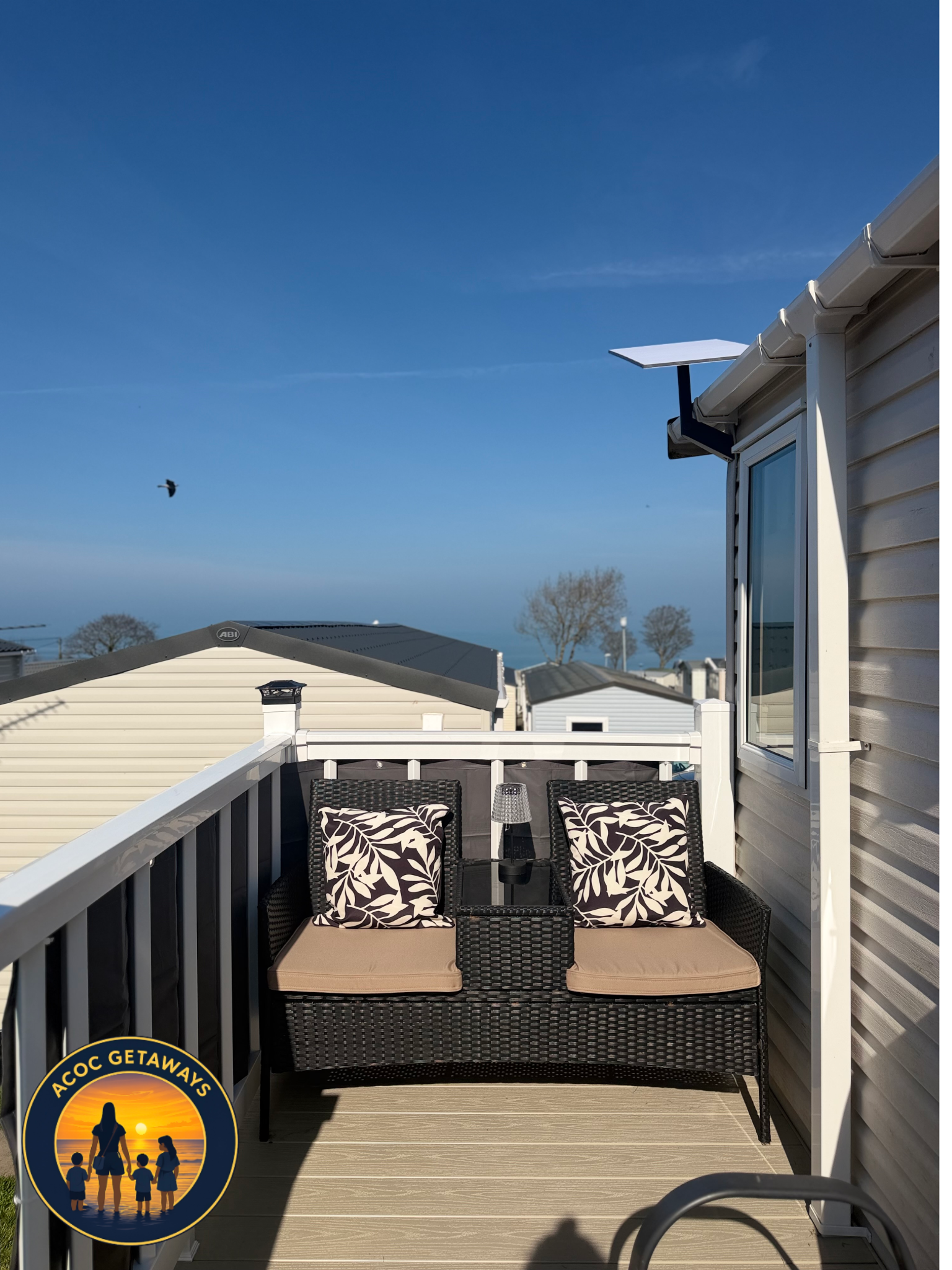 A balcony with a black wicker sofa, two matching cushions with white and black leaf patterns, a small black side table with a glass top, and a beige cushion. The balcony has a white railing and is attached to a house with beige siding. The sky is blu