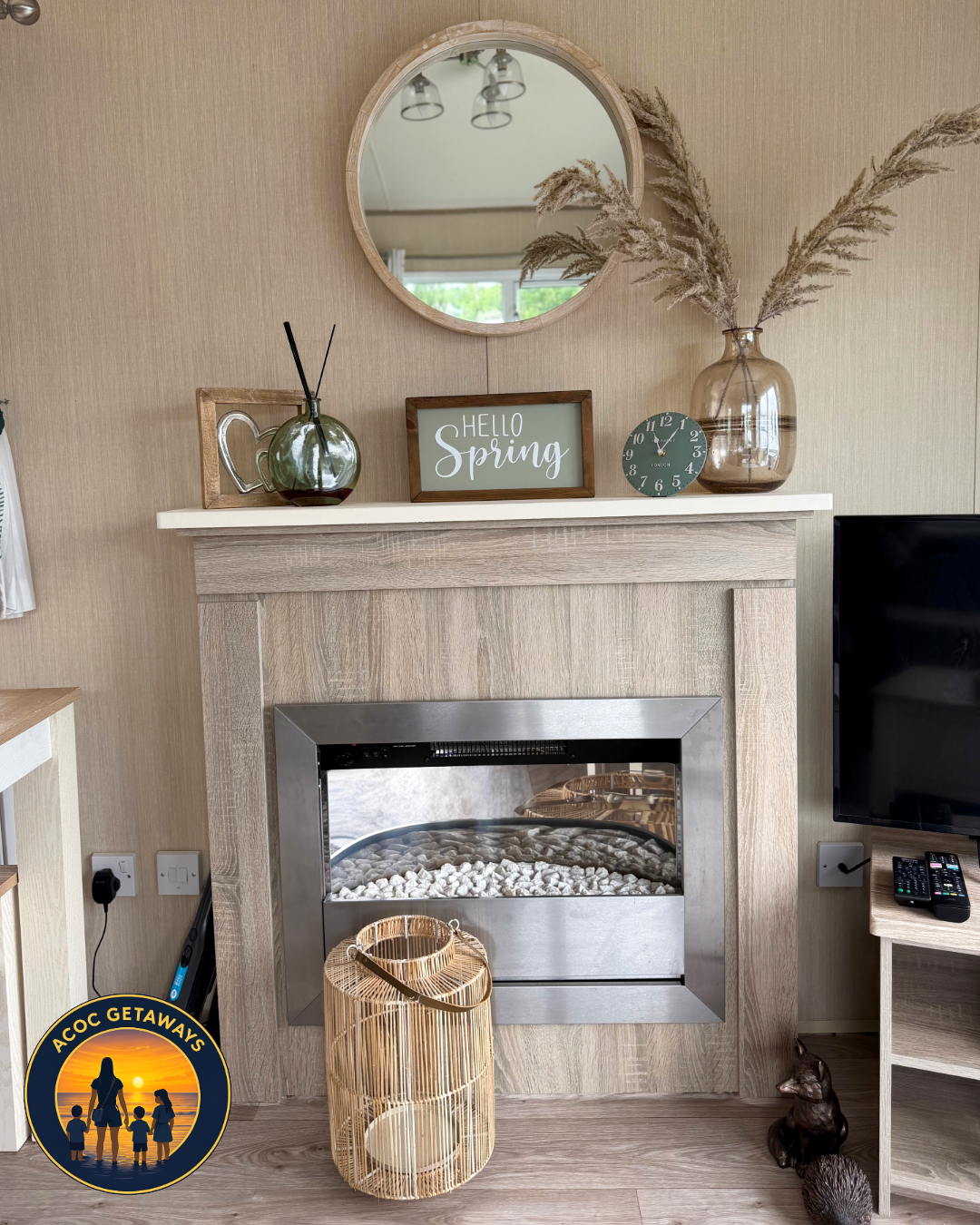 Living room fireplace with a round mirror above and a shelf decorated with a green glass vase with pampas grass, a framed 'Hello Spring' sign, a green clock, and a small picture frame. There's a large wicker lantern in front of the fireplace, a telev