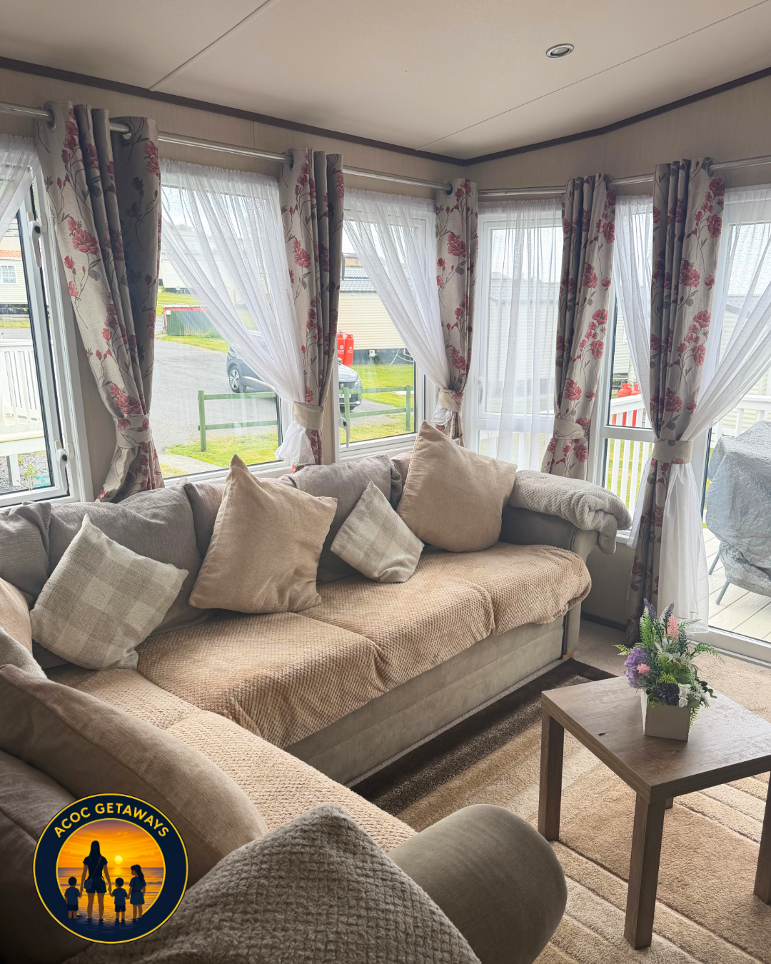 Living room with beige sofa and multiple pillows, surrounding windows with floral curtains and sheer white drapes, wooden coffee table with a flower arrangement, view of outdoors with parked car and a deck.